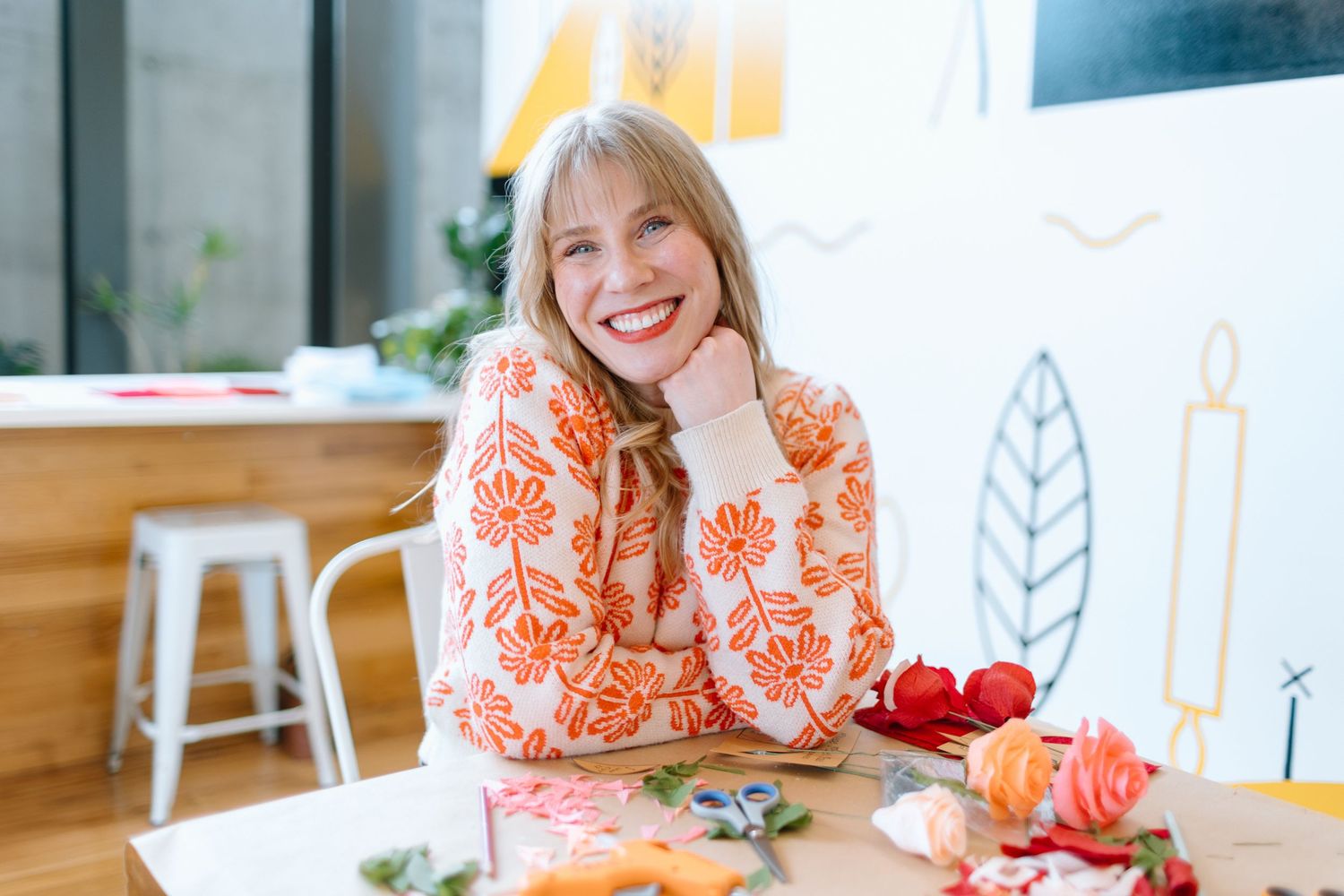 Smiling blonde woman in orange floral blouse sitting at a table with flowers in a bright modern studio.