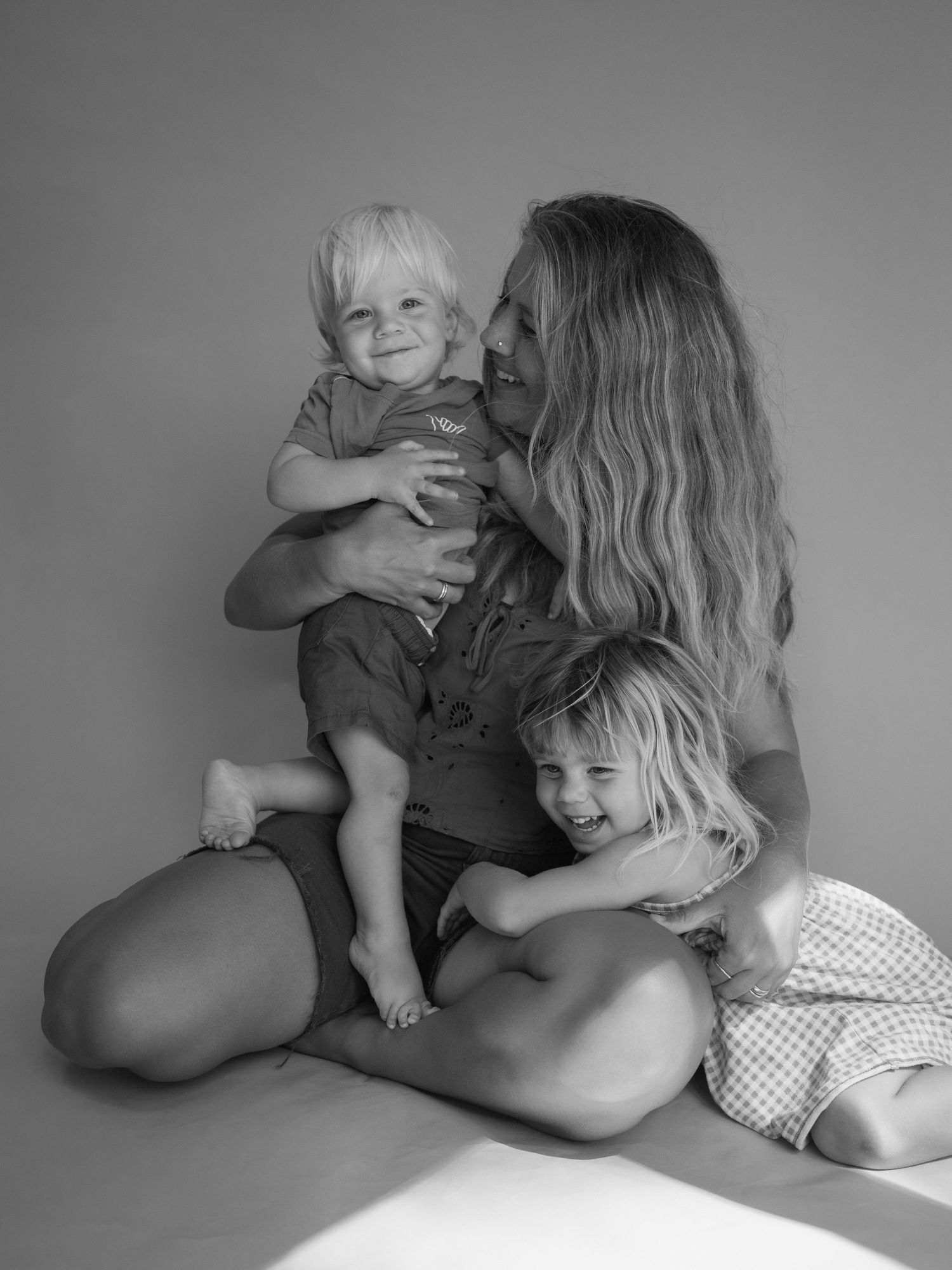 Black and white photo of a mother sitting on the floor, lovingly holding two young blonde children.