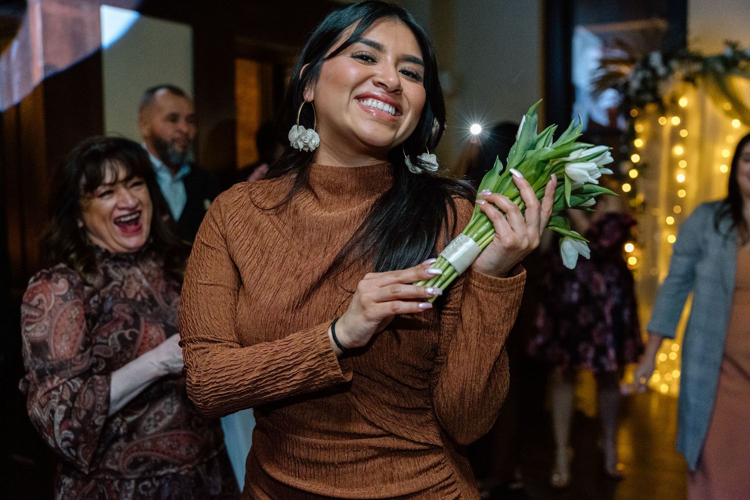Woman in brown dress smiling while catching a bridal bouquet of white tulips at a wedding reception.