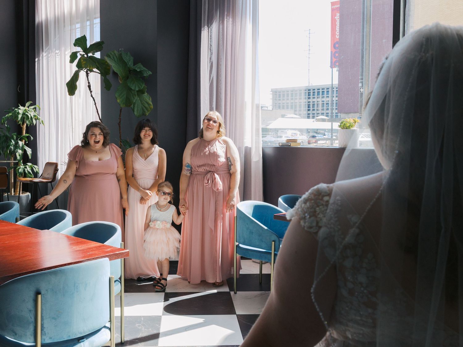 Bridesmaids in pink gowns react with joy as bride in white wedding dress and veil reveals her look.