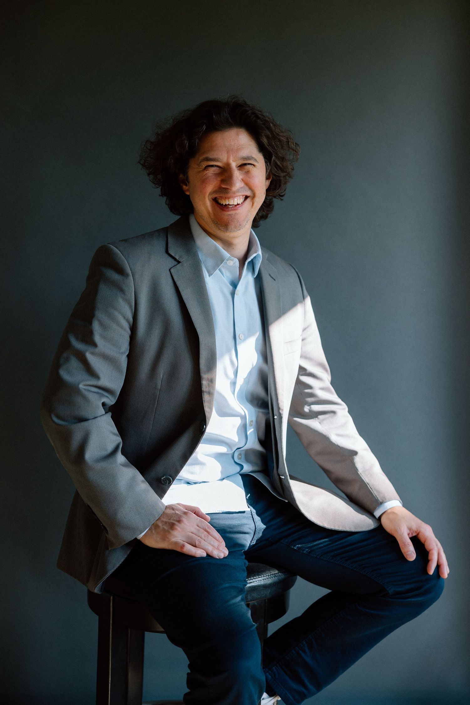 Smiling man with curly hair in gray blazer and blue shirt sits relaxed on stool against dark background.
