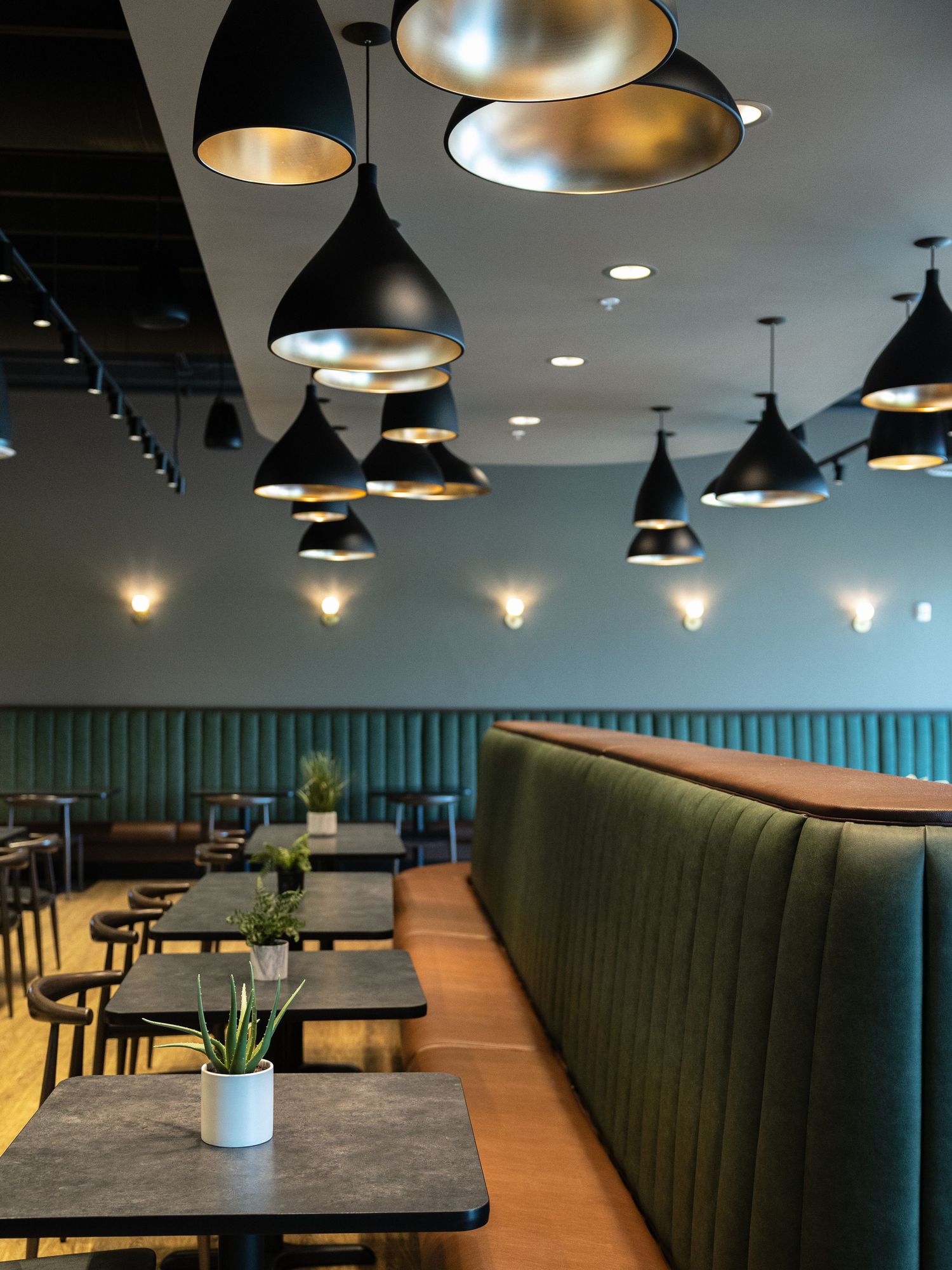 Modern restaurant interior with black pendant lights, green velvet booth seating, and small potted plants on dark tables.