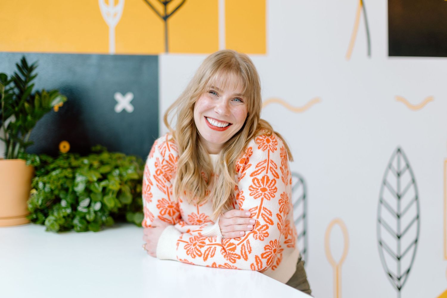 Smiling blonde woman in orange floral dress seated at white table with green plants and colorful wall decor.