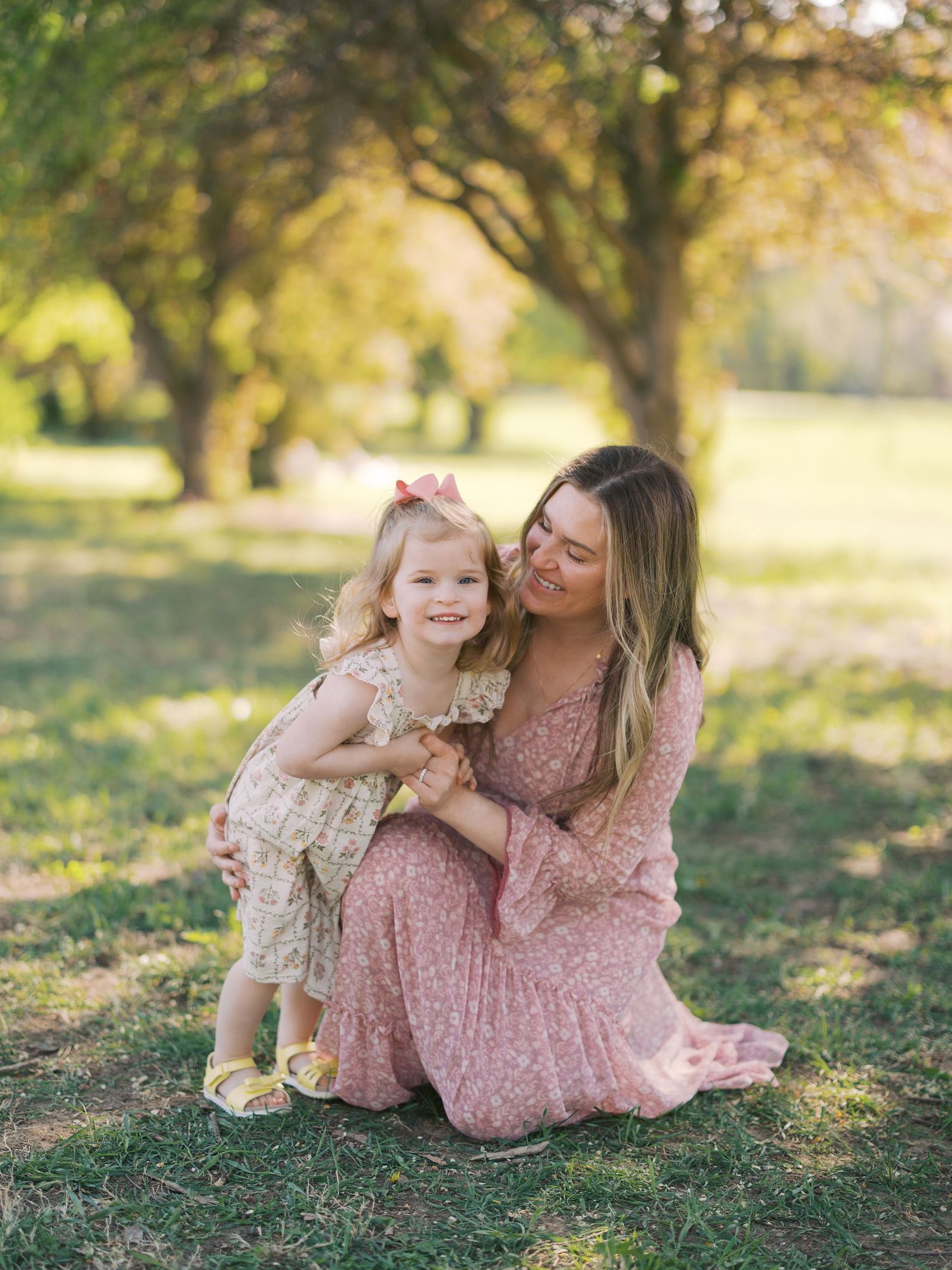 A mother in a pink floral dress kneels on grass, smiling at her toddler daughter in a sunny park.