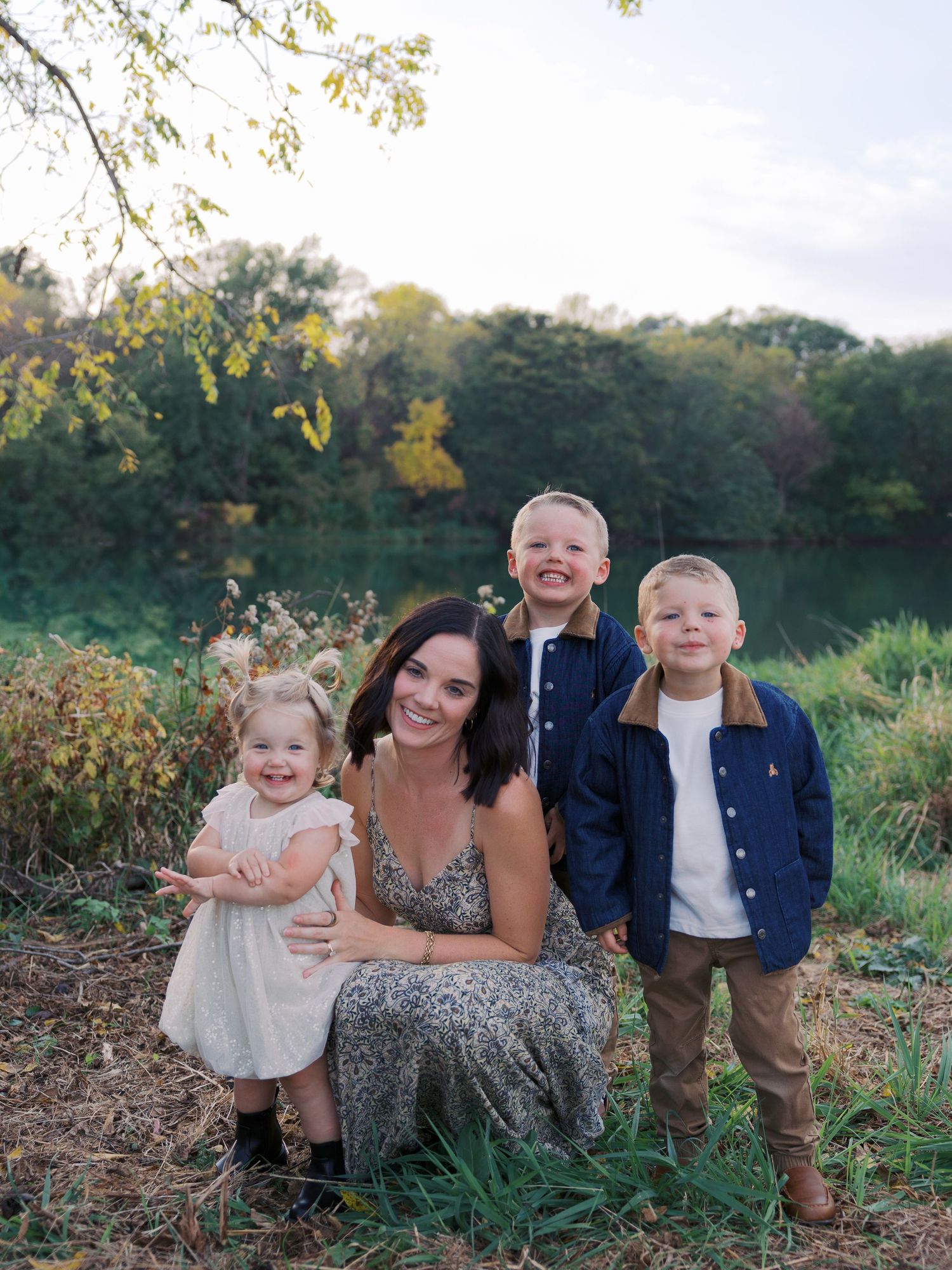 A smiling mother poses outdoors with her three young children near a serene pond surrounded by lush green trees.