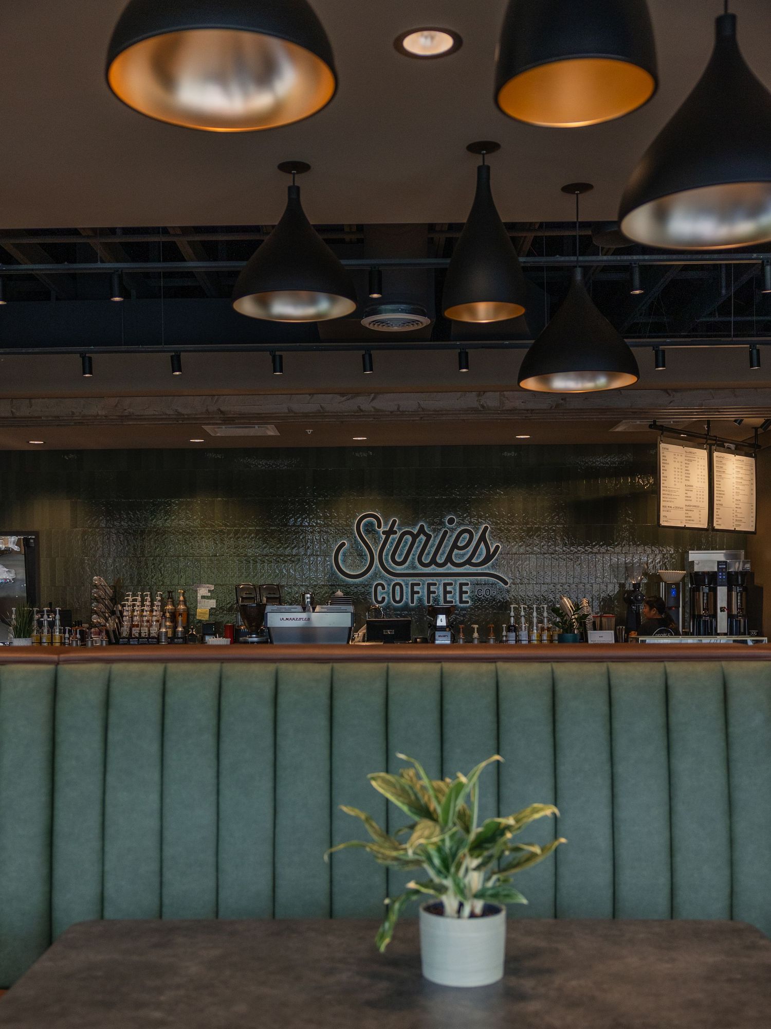 Interior of Stories Coffee shop featuring a green tufted booth, pendant lights, and a neon sign above the bar.