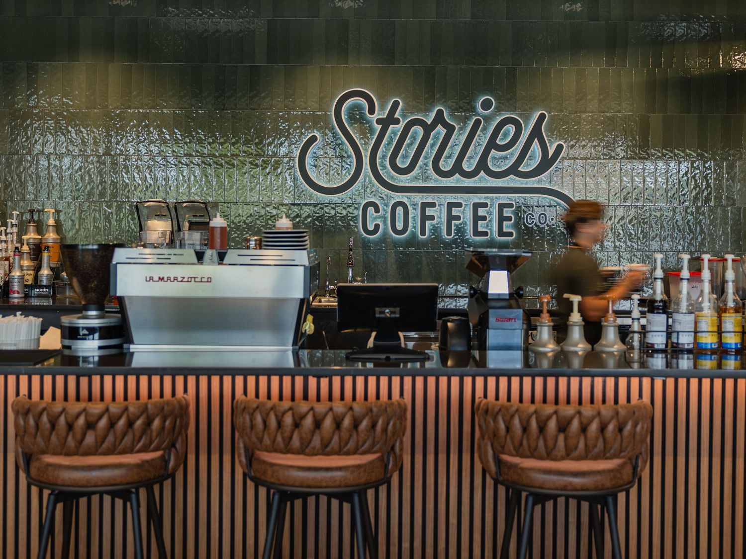 Stories Coffee bar interior with espresso machine, leather stools, and neon sign above a wood-paneled counter.