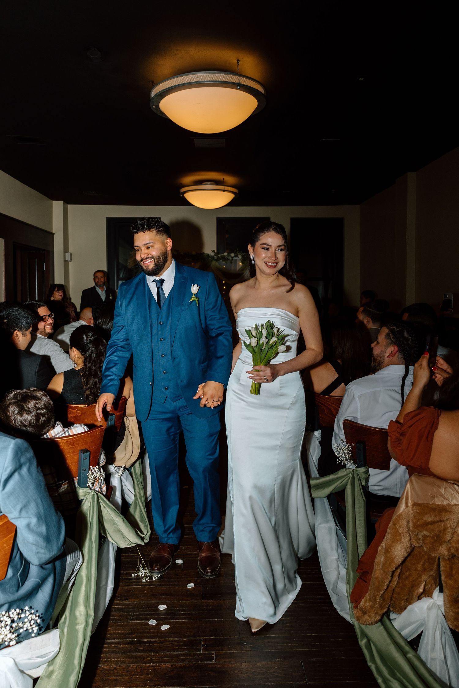 Bride in white gown holding tulips walks with groom in blue suit down wedding aisle as guests look on.