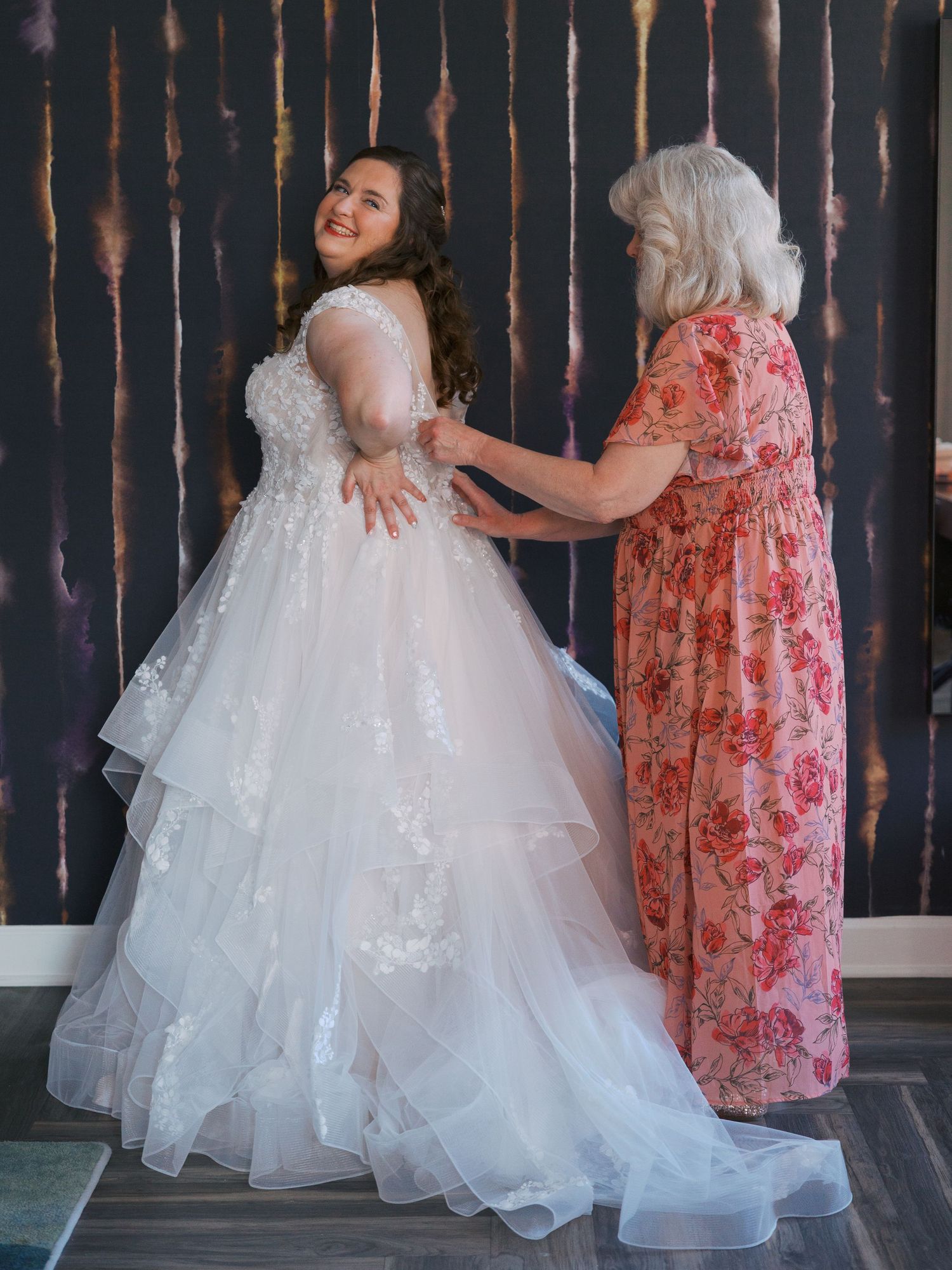 Bride in layered white lace ball gown being helped by older woman in floral dress before wedding ceremony.