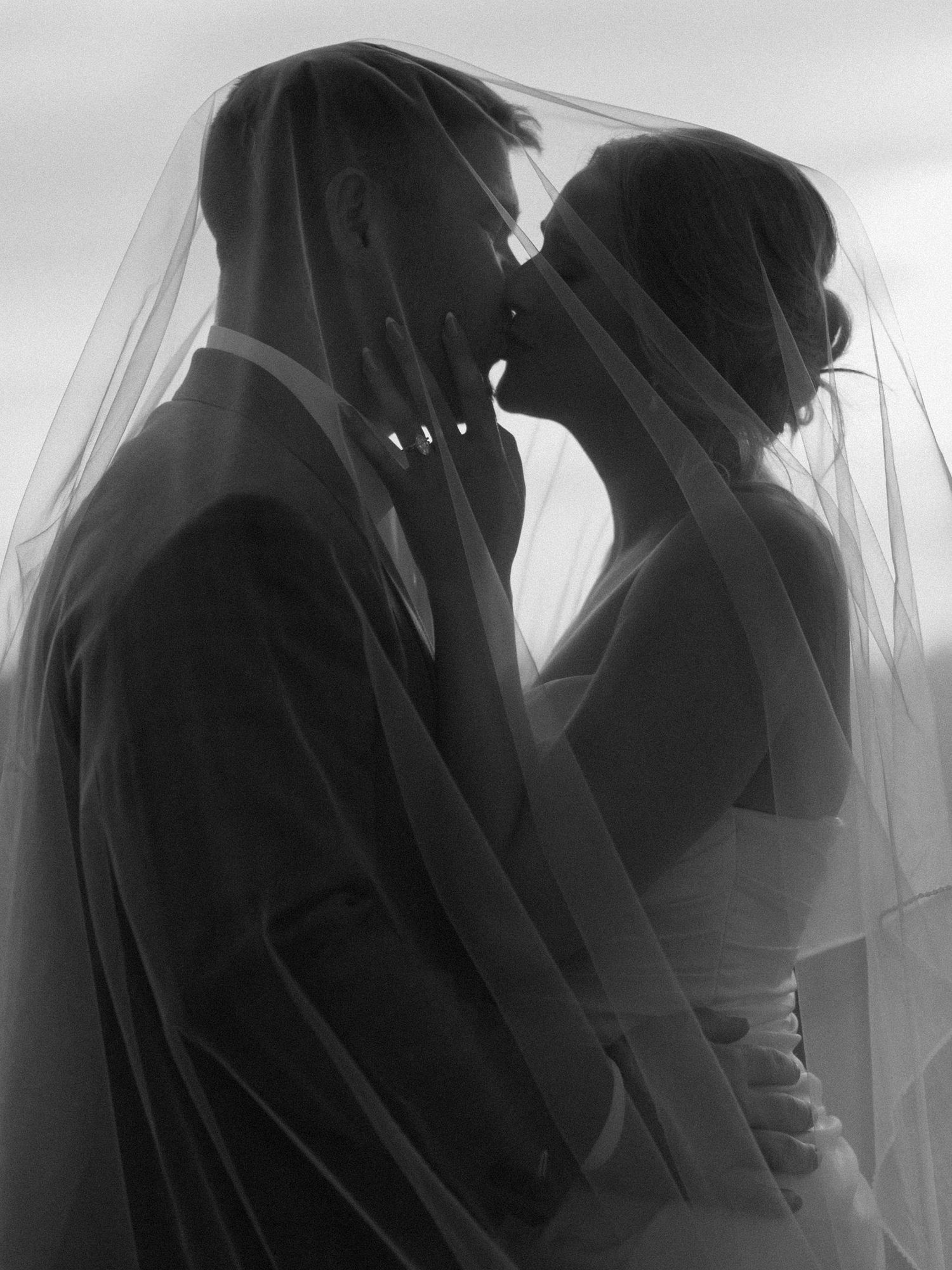 Black and white photo of bride and groom sharing a kiss under a flowing wedding veil.