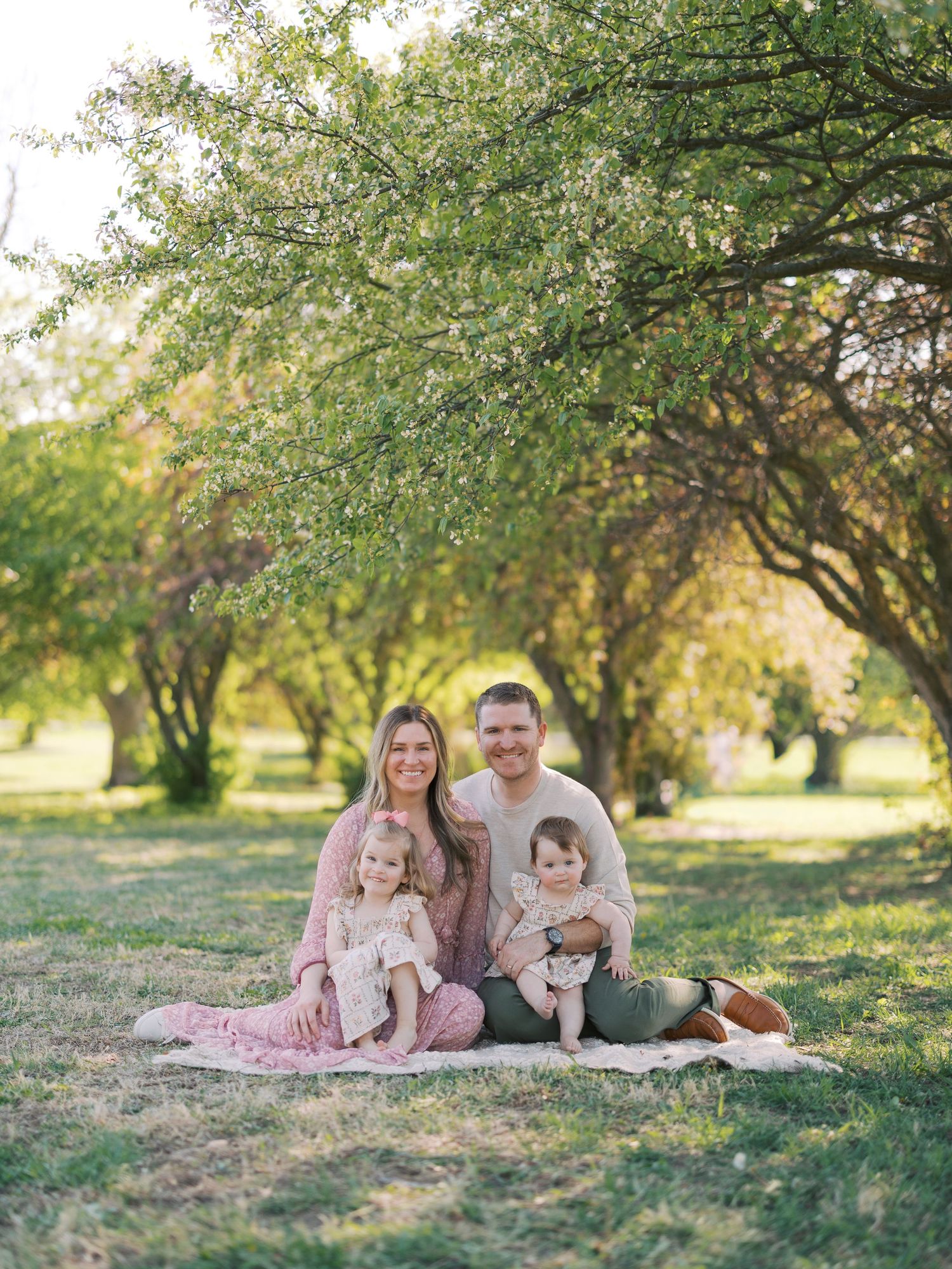 Family of four sitting on a blanket under lush green trees in a park, smiling warmly during a spring outdoor photo session.