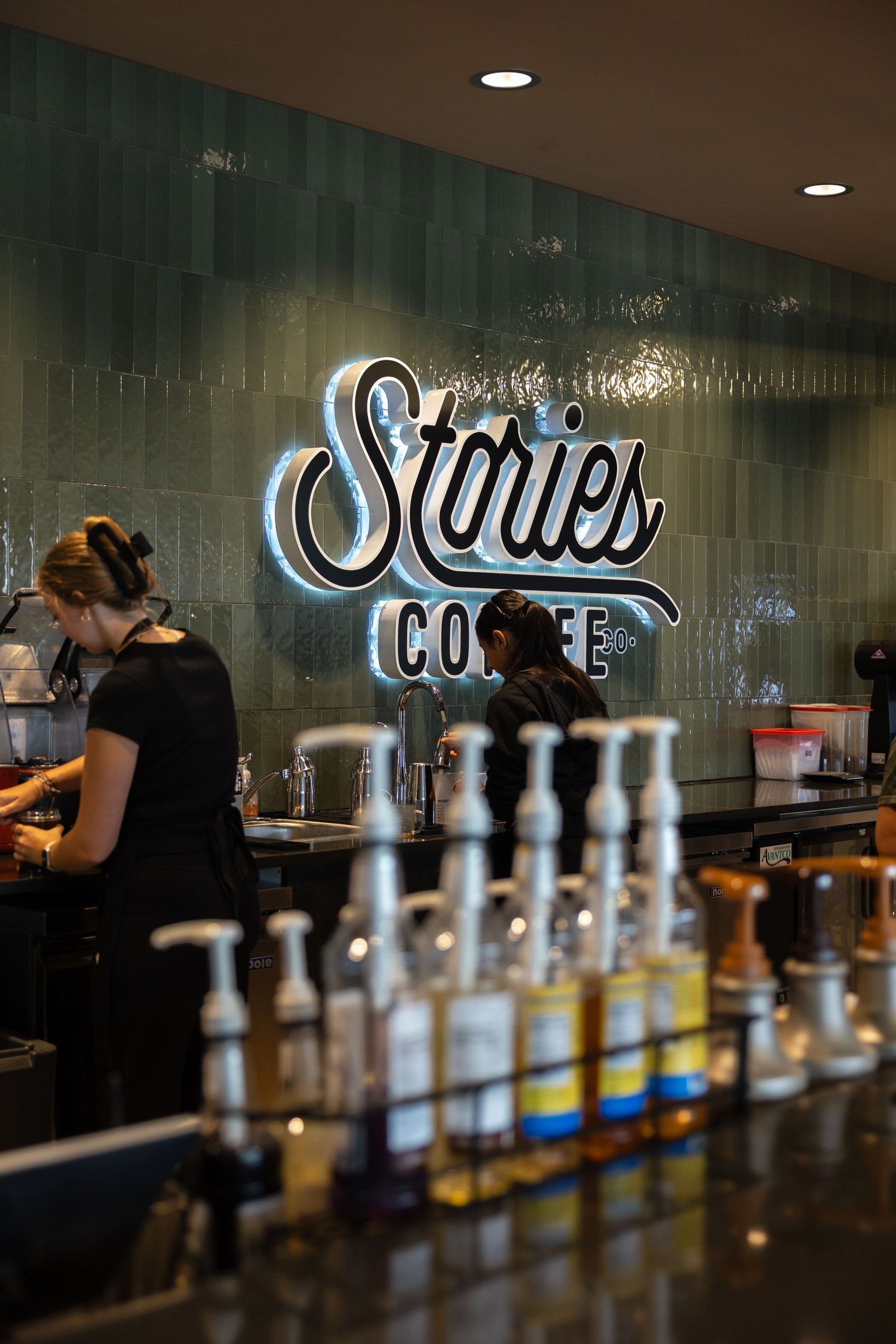 Barista working behind the counter at Stories Coffee shop with syrup bottles in foreground and glowing sign on tiled wall.