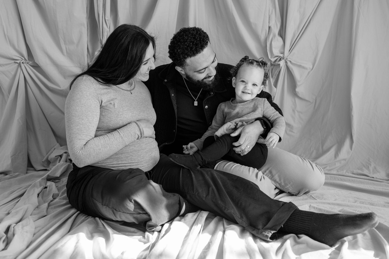 Black and white family portrait of parents sitting on fabric backdrop with their smiling toddler.