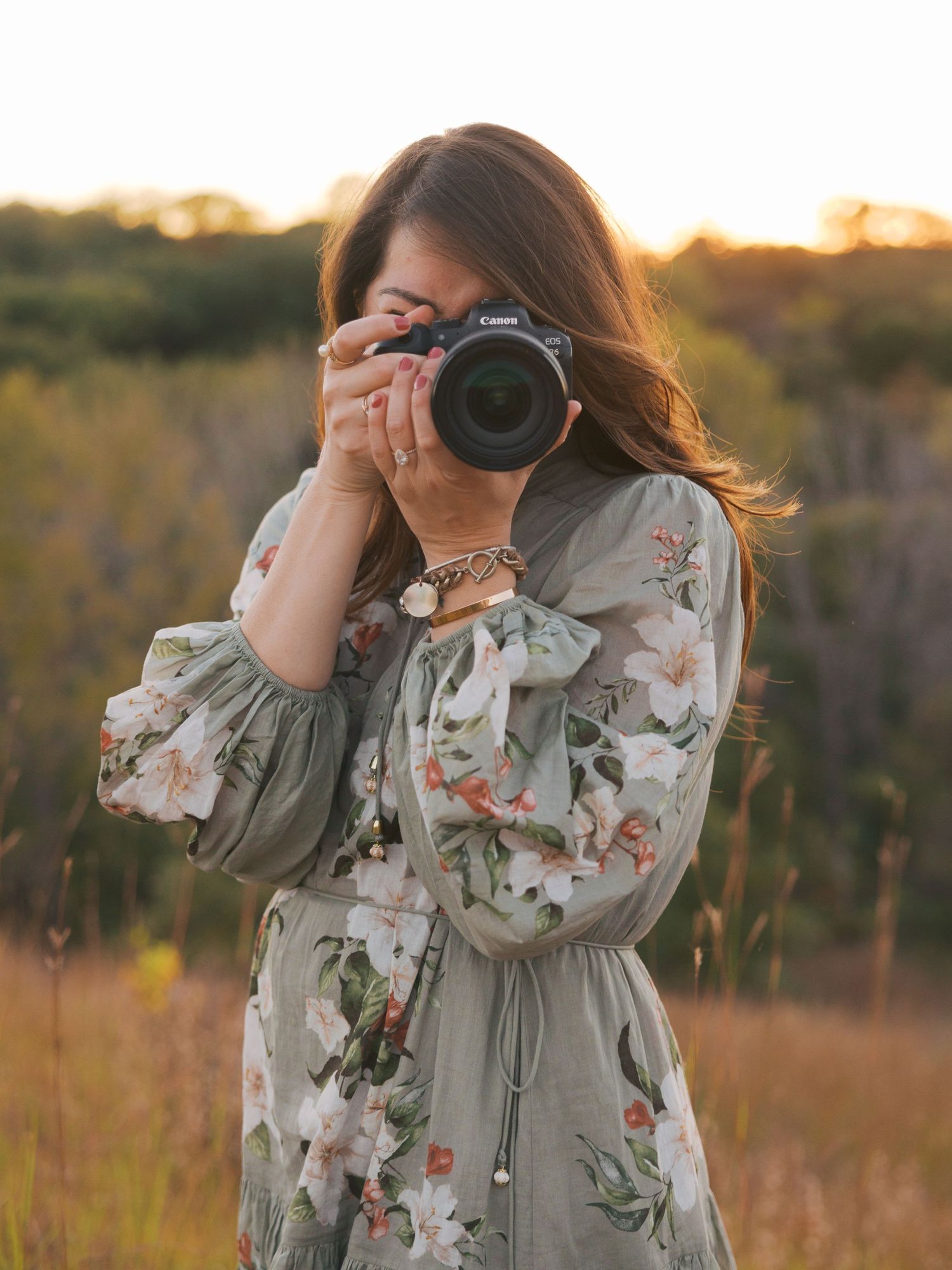 Woman in floral dress photographing outdoors with Canon camera at golden hour in a natural field setting.