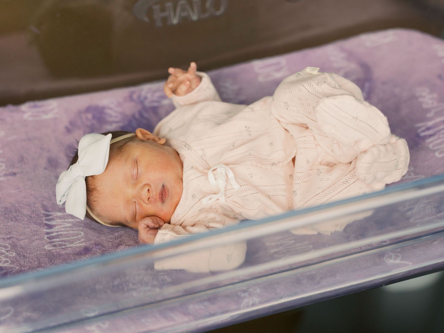Newborn baby girl sleeping peacefully in hospital bassinet, wearing pink outfit and white bow headband.