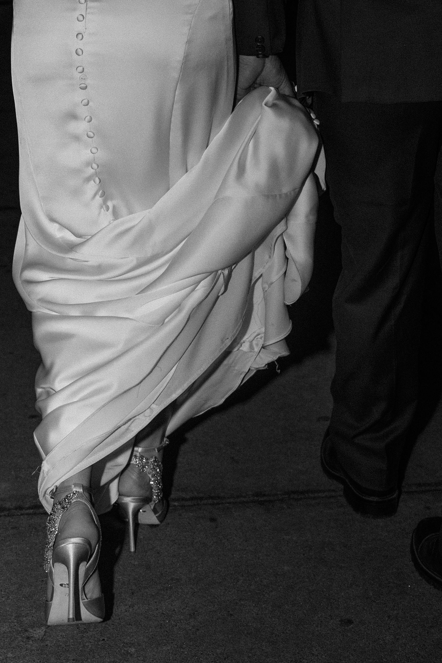 Black and white photo of a bride in a button-back gown and heels walking with a groom.