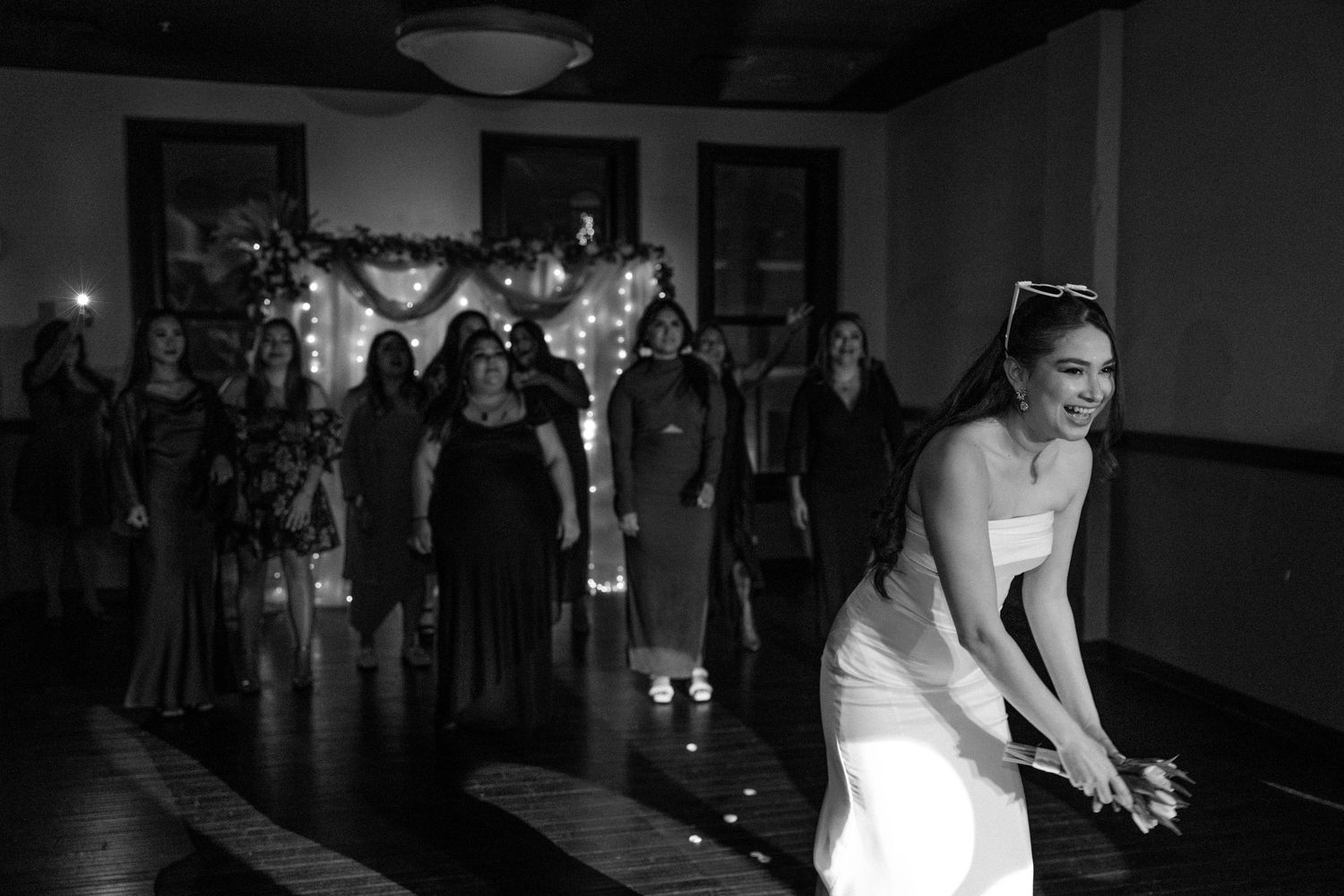 Black and white photo of a bride tossing her bouquet to a group of women at a wedding reception.