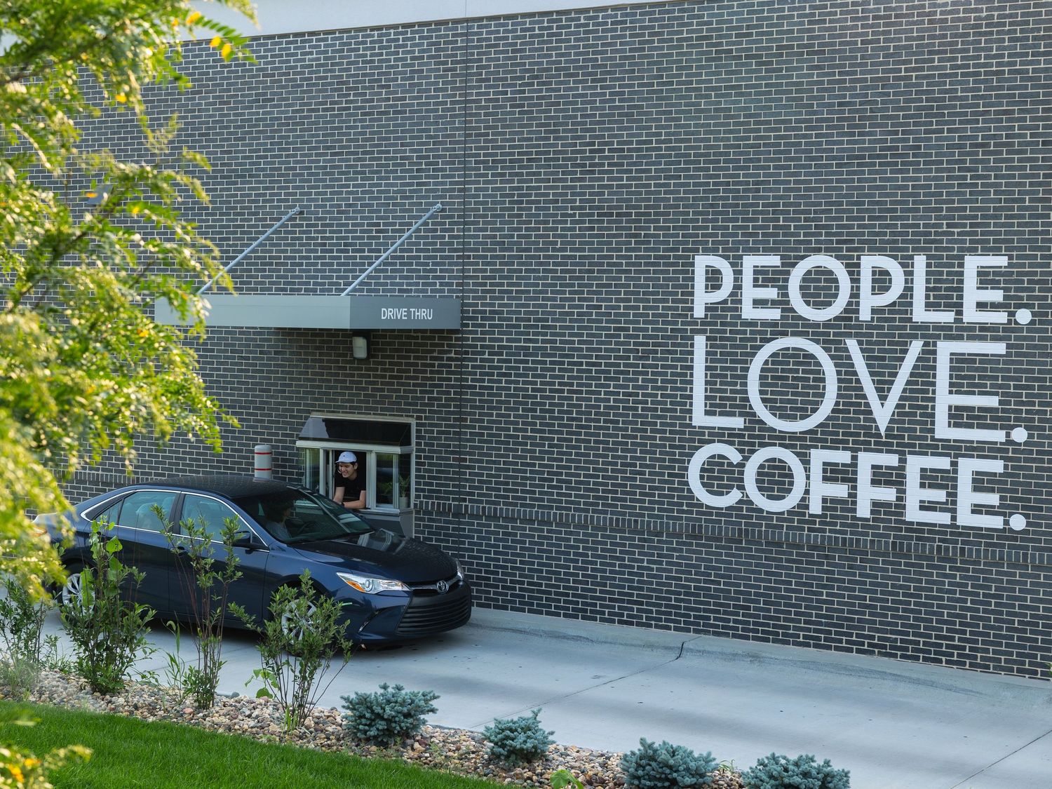 Modern brick building exterior with bold white text reading People Love Coffee on the wall, with a car parked outside.
