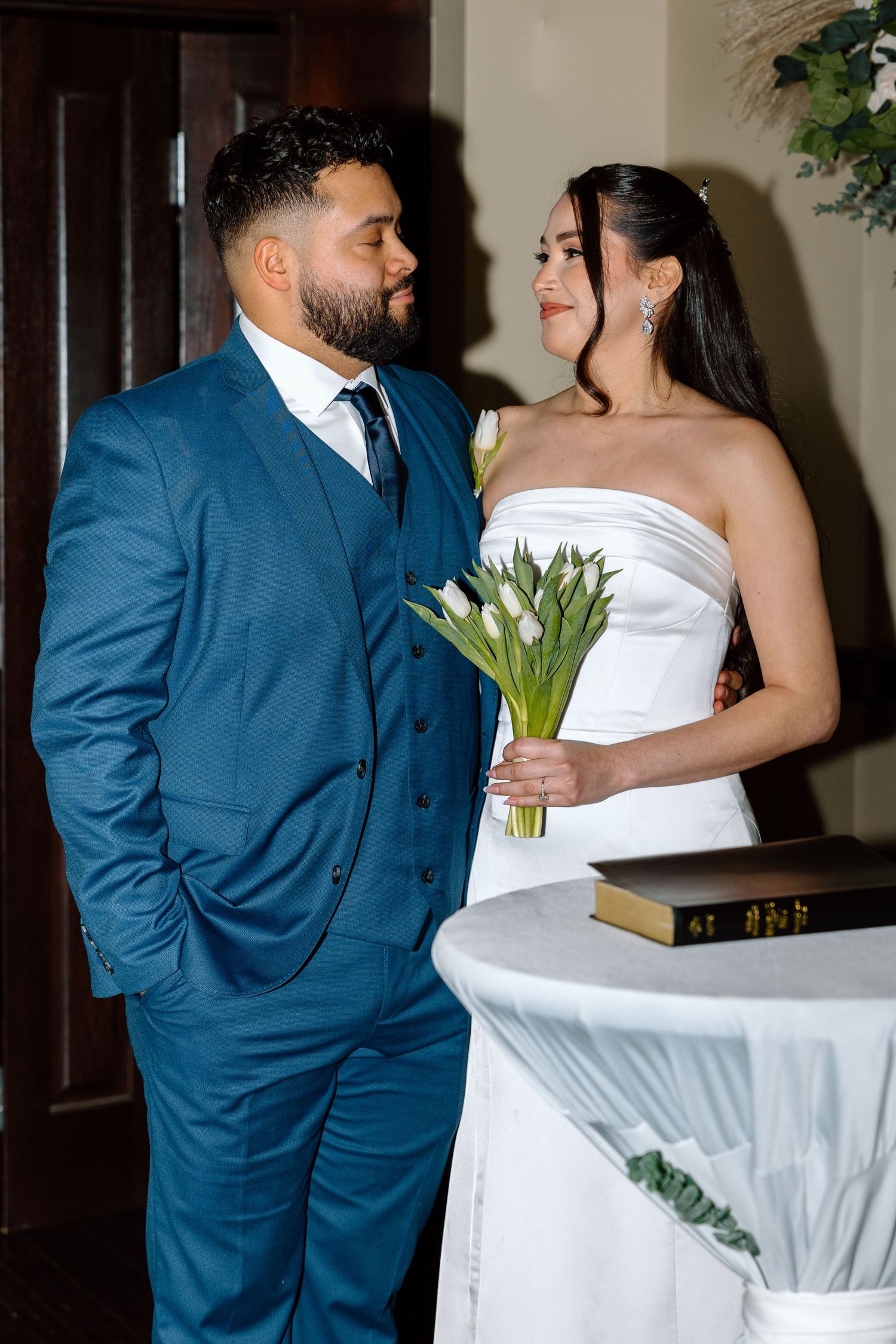 A groom in a blue suit and bride in a white strapless gown holding white tulips share a loving glance at their wedding.