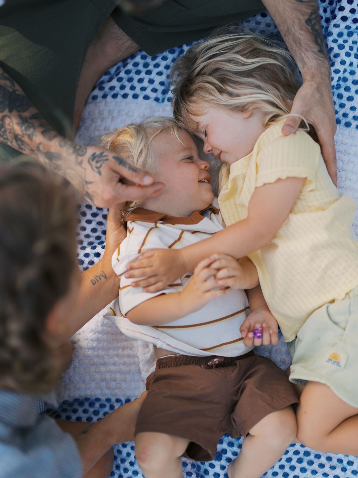 Two toddlers lying on a polka dot blanket, touching foreheads and smiling, surrounded by adults outdoors.