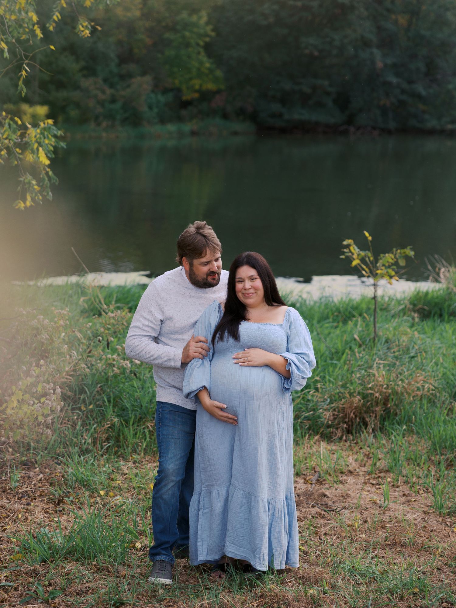 A couple poses by a serene lake, the woman in a blue maternity dress cradling her baby bump during an outdoor maternity photo session.