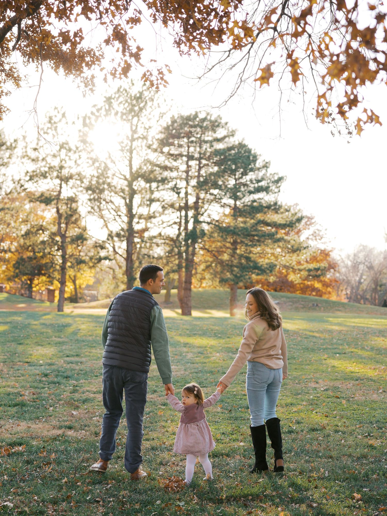 A family of three holds hands in a sunlit autumn park, parents swinging their toddler between them.