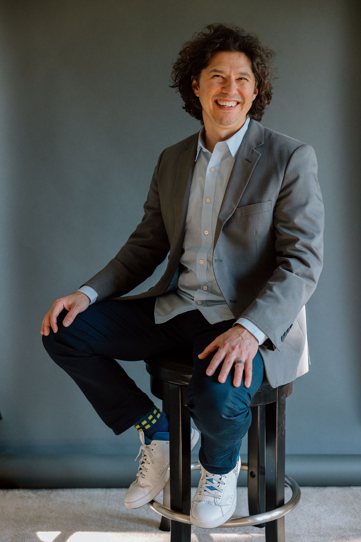 Smiling man with curly hair sits on stool wearing gray blazer, jeans, and white sneakers against gray background.