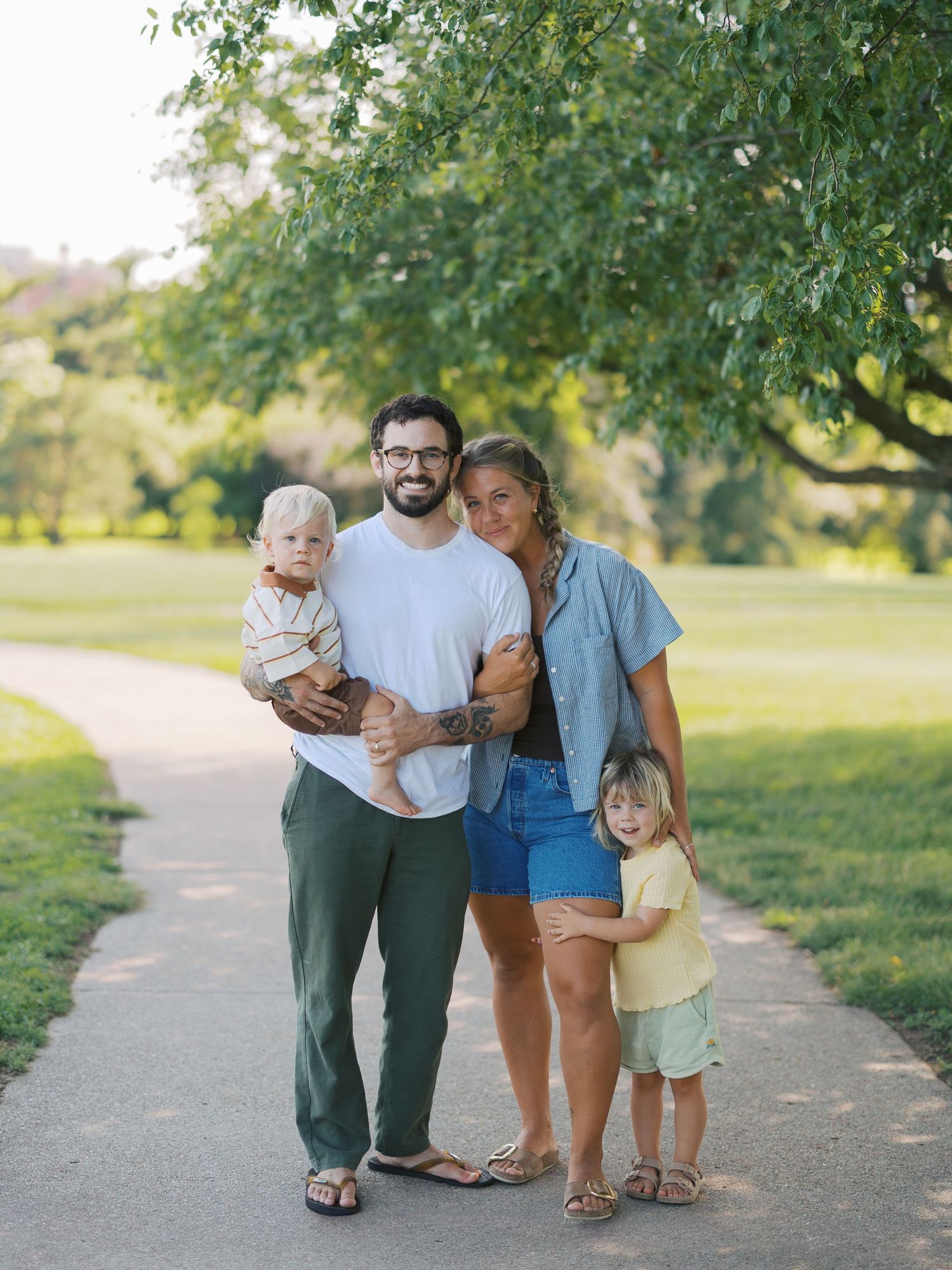 Family of four posing on a park path, parents holding young children, surrounded by lush green trees on a sunny day.