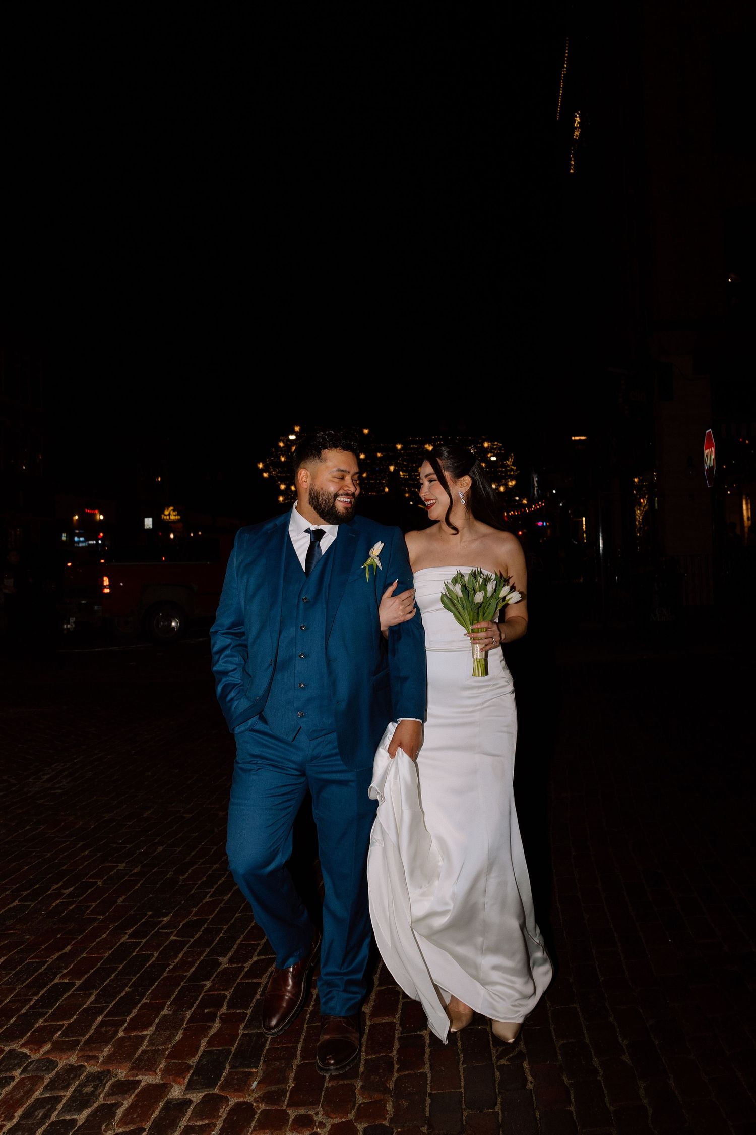 Bride and groom walking together at night, she holds green tulips in white gown, he wears a navy blue suit.