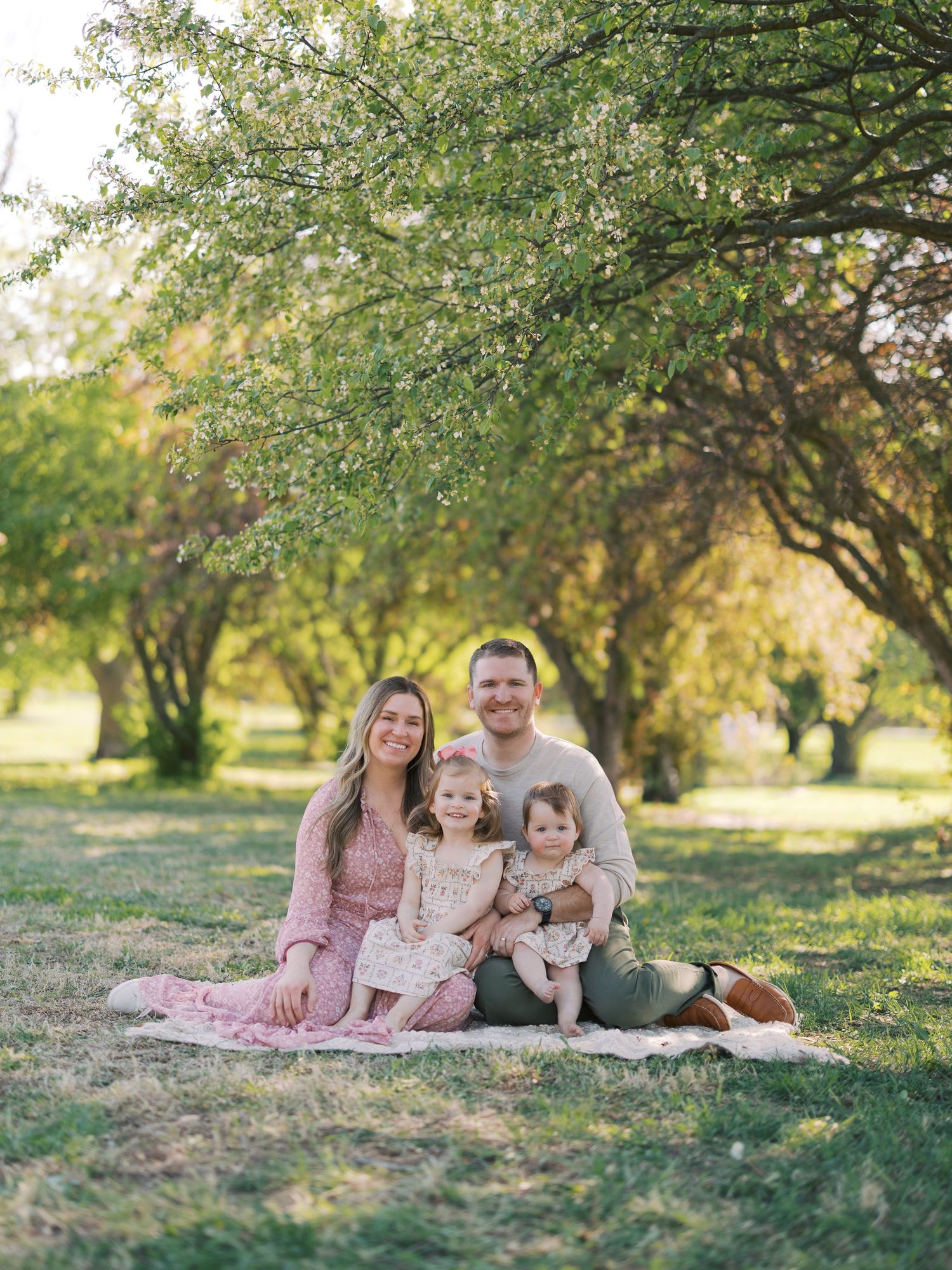 Family of four sitting on a blanket in a sunlit park with blooming trees during a spring outdoor photo session.