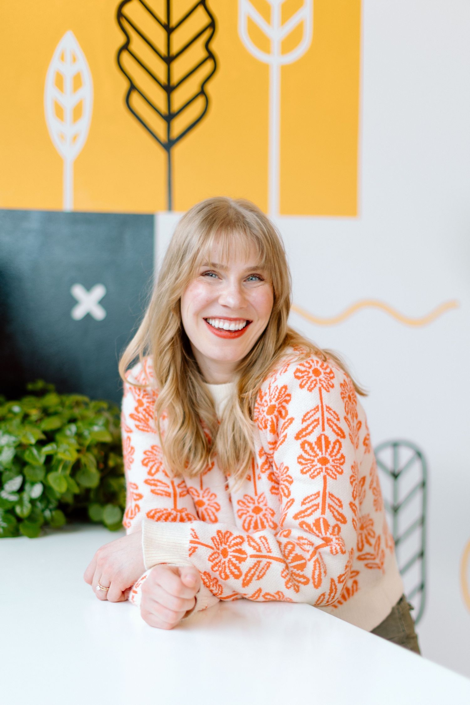 Smiling blonde woman in orange floral sweater seated at white table with yellow botanical wall art background.