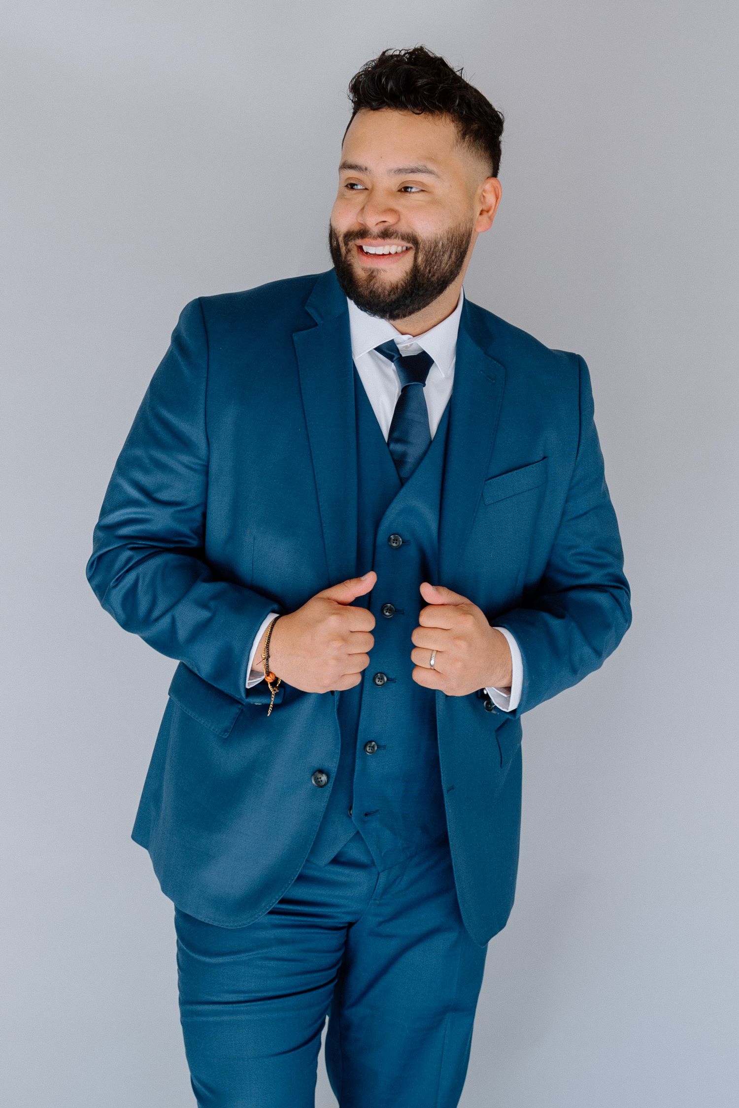 Smiling man in a blue three-piece suit with tie, posing confidently against a gray background.
