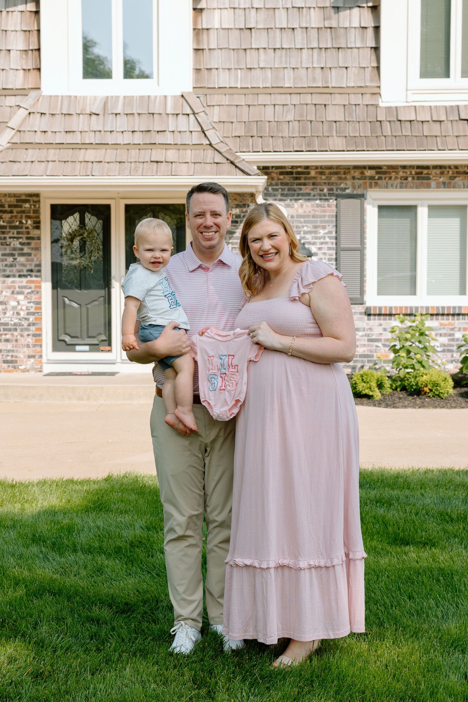 Happy family of three posing outdoors, pregnant woman in pink dress holding baby onesie, man holding toddler, brick house background.