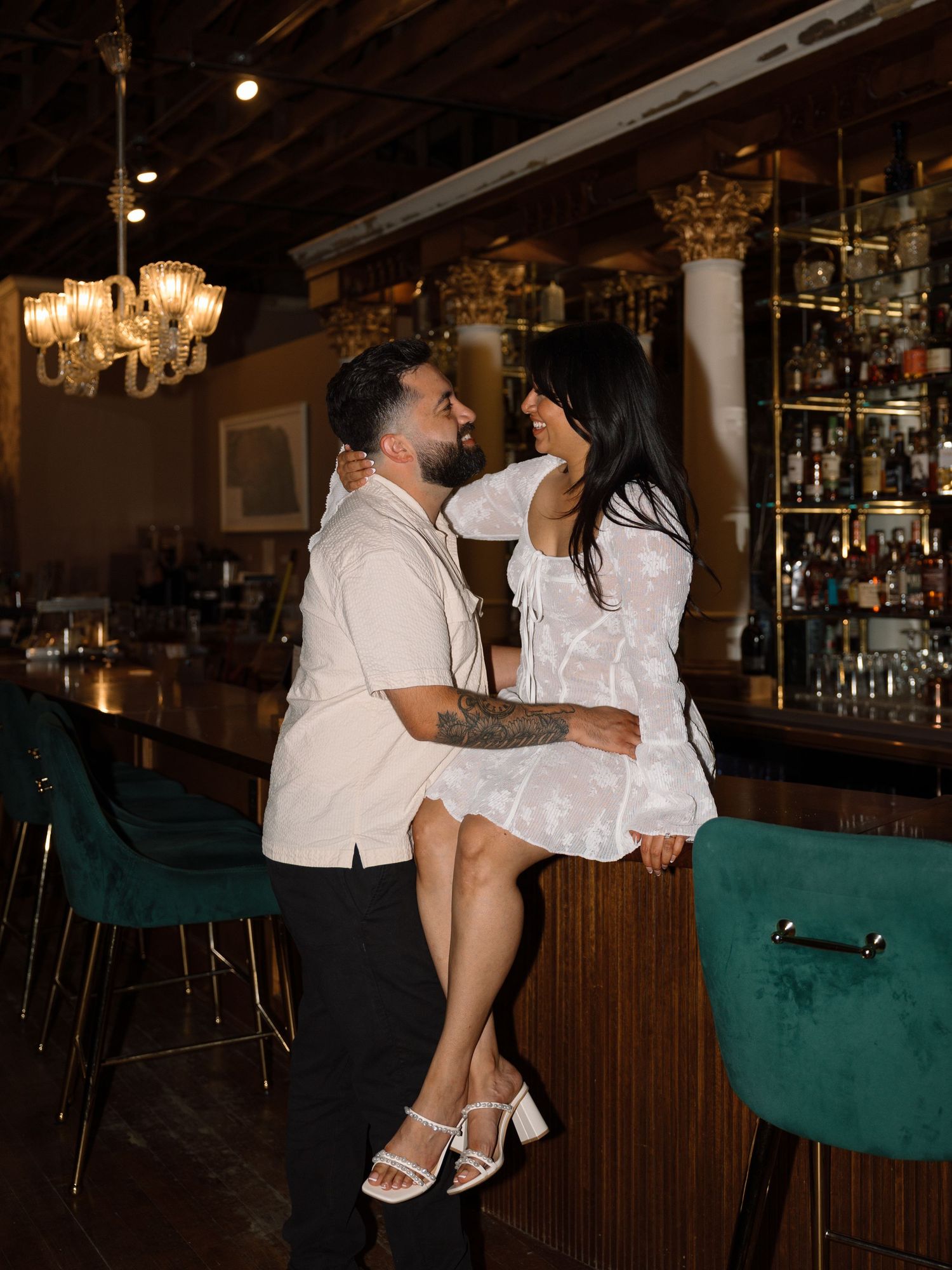Couple sharing a romantic moment at an elegant bar with green velvet stools and chandelier lighting.