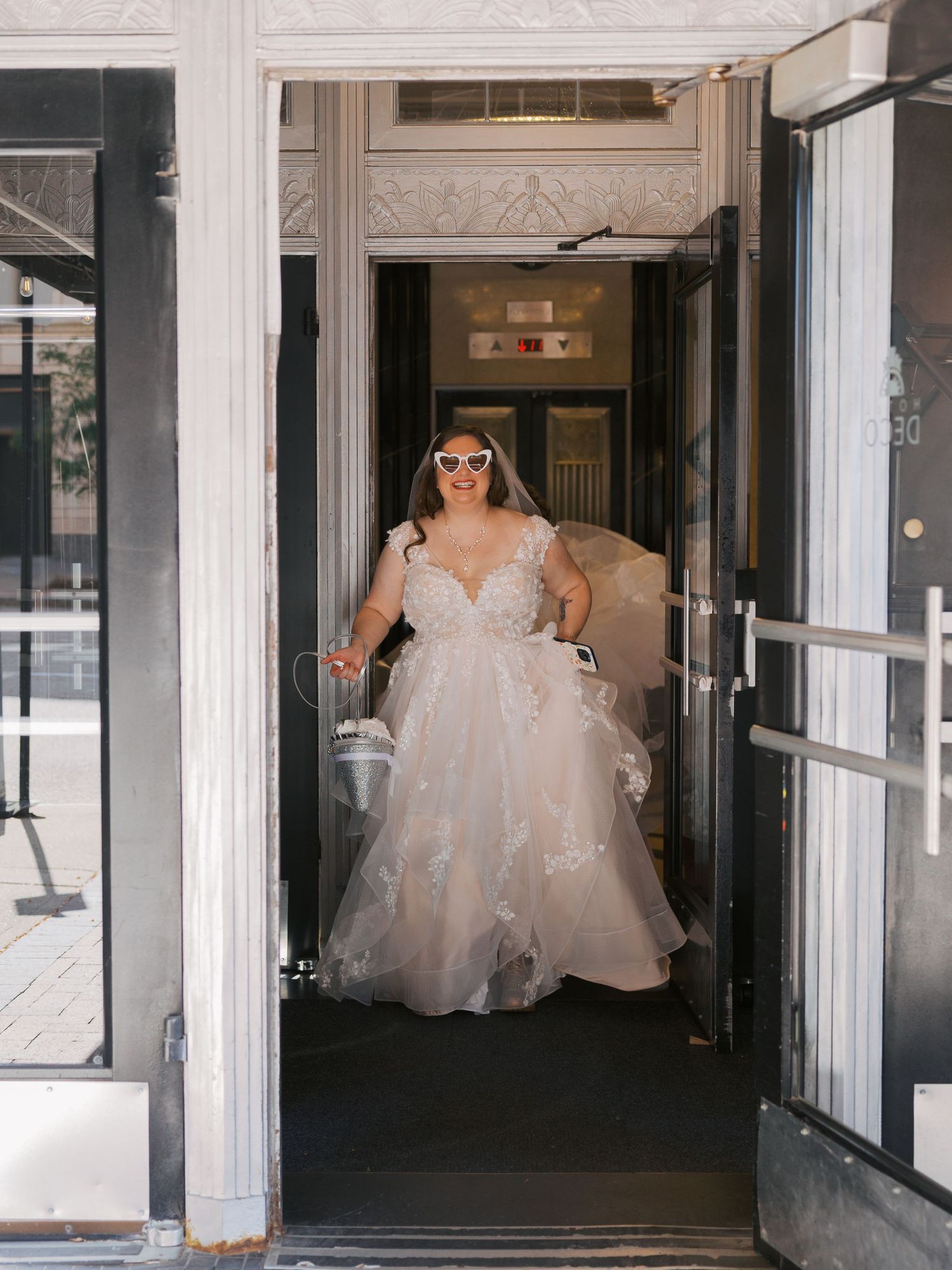 Smiling bride in a lace wedding gown and white sunglasses walking through a doorway carrying a flower girl basket.