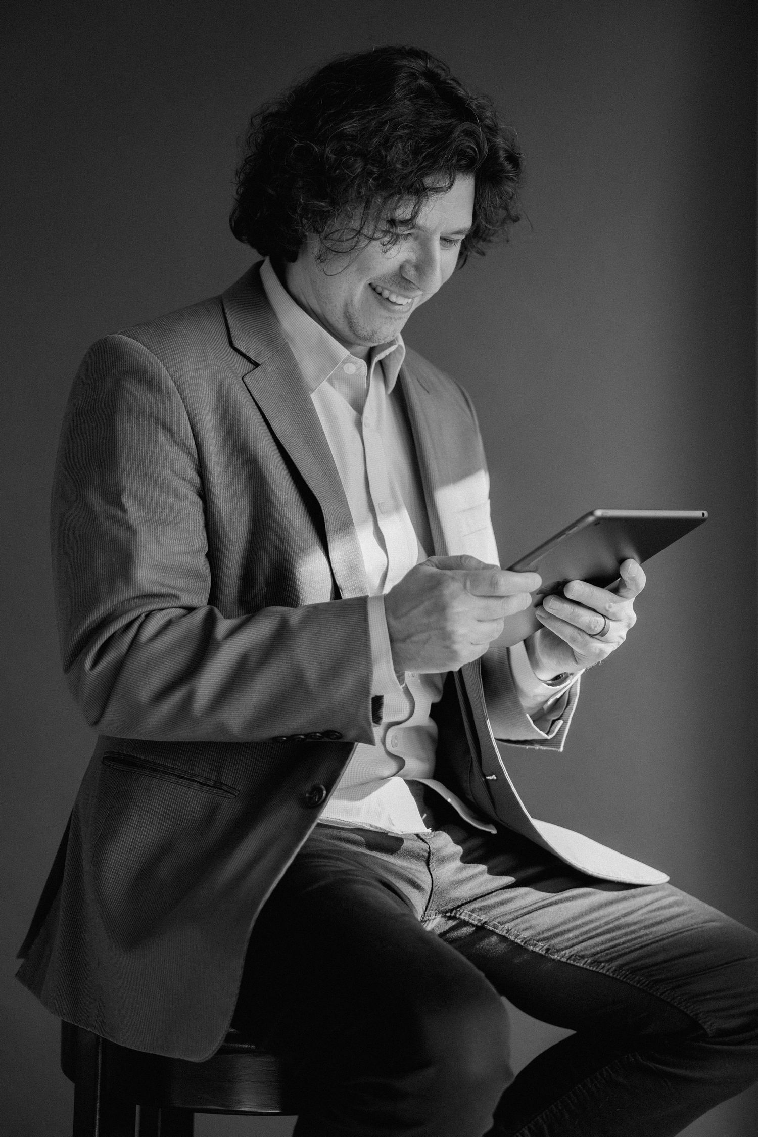 Black and white photo of a smiling man in a blazer sitting and using a digital tablet.