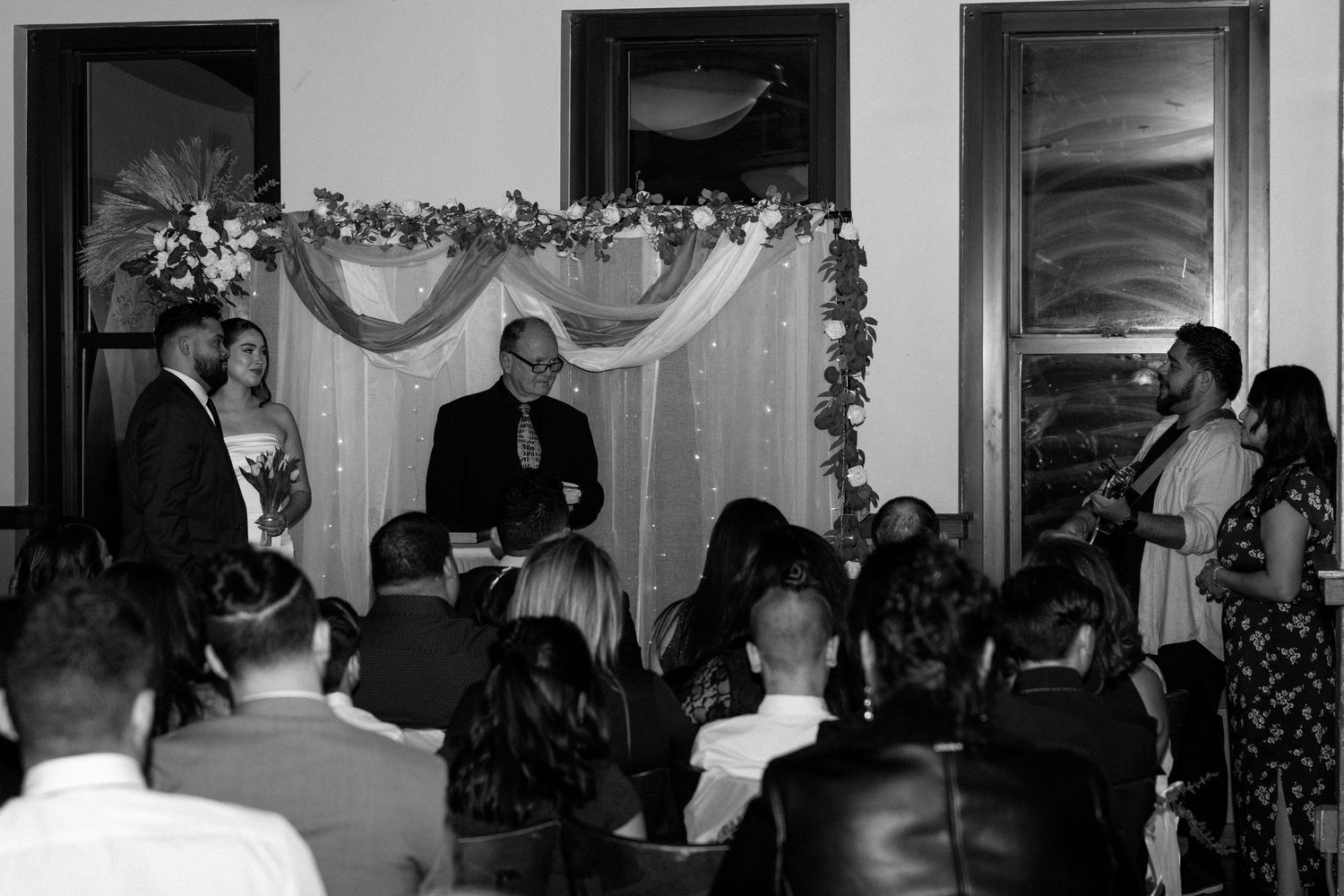 Black and white wedding ceremony photo with couple at altar, officiant, and seated guests in elegant indoor venue.