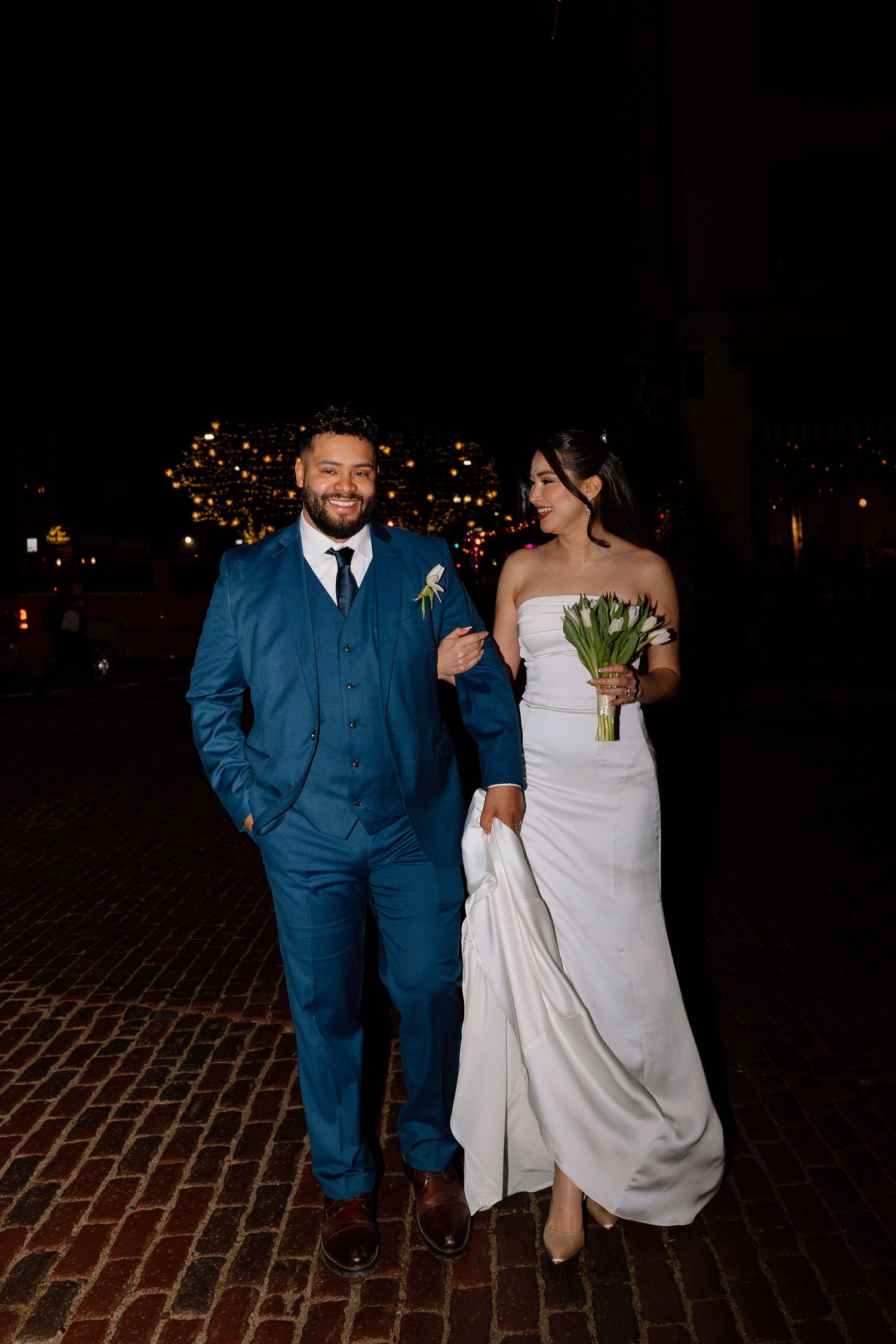 Couple walking together at night on brick pavement, bride in white dress and groom in blue suit.