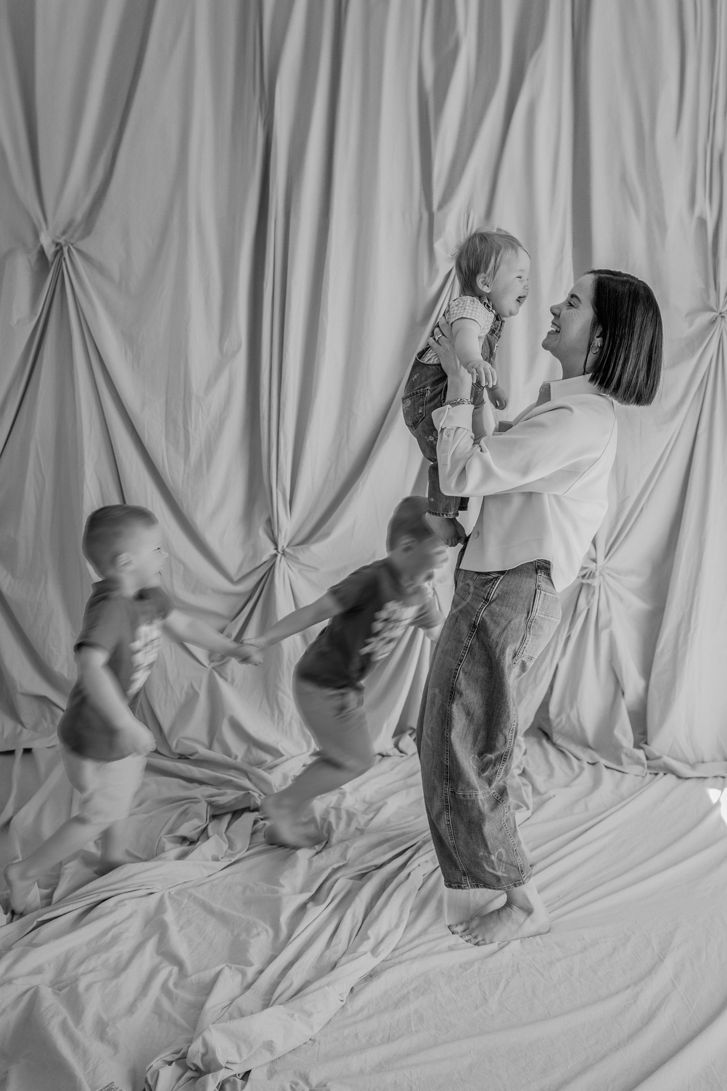 Playful black and white studio portrait with children jumping on draped fabric backdrop.