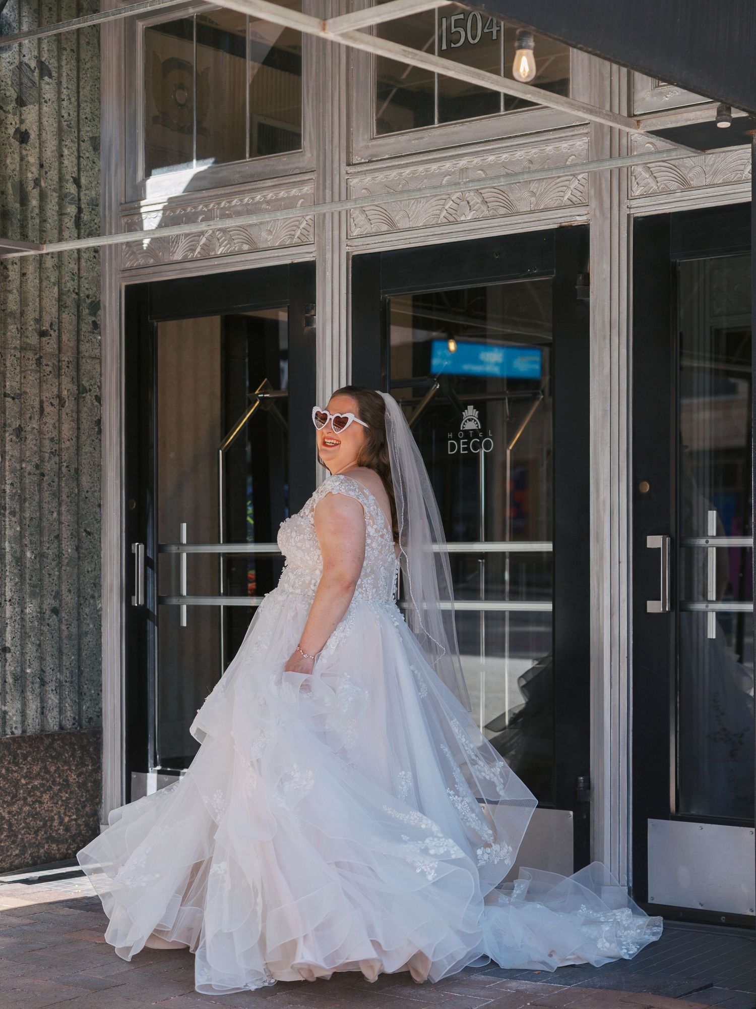 Bride in white lace ball gown and veil poses outside building 150, wearing white sunglasses on her wedding day.