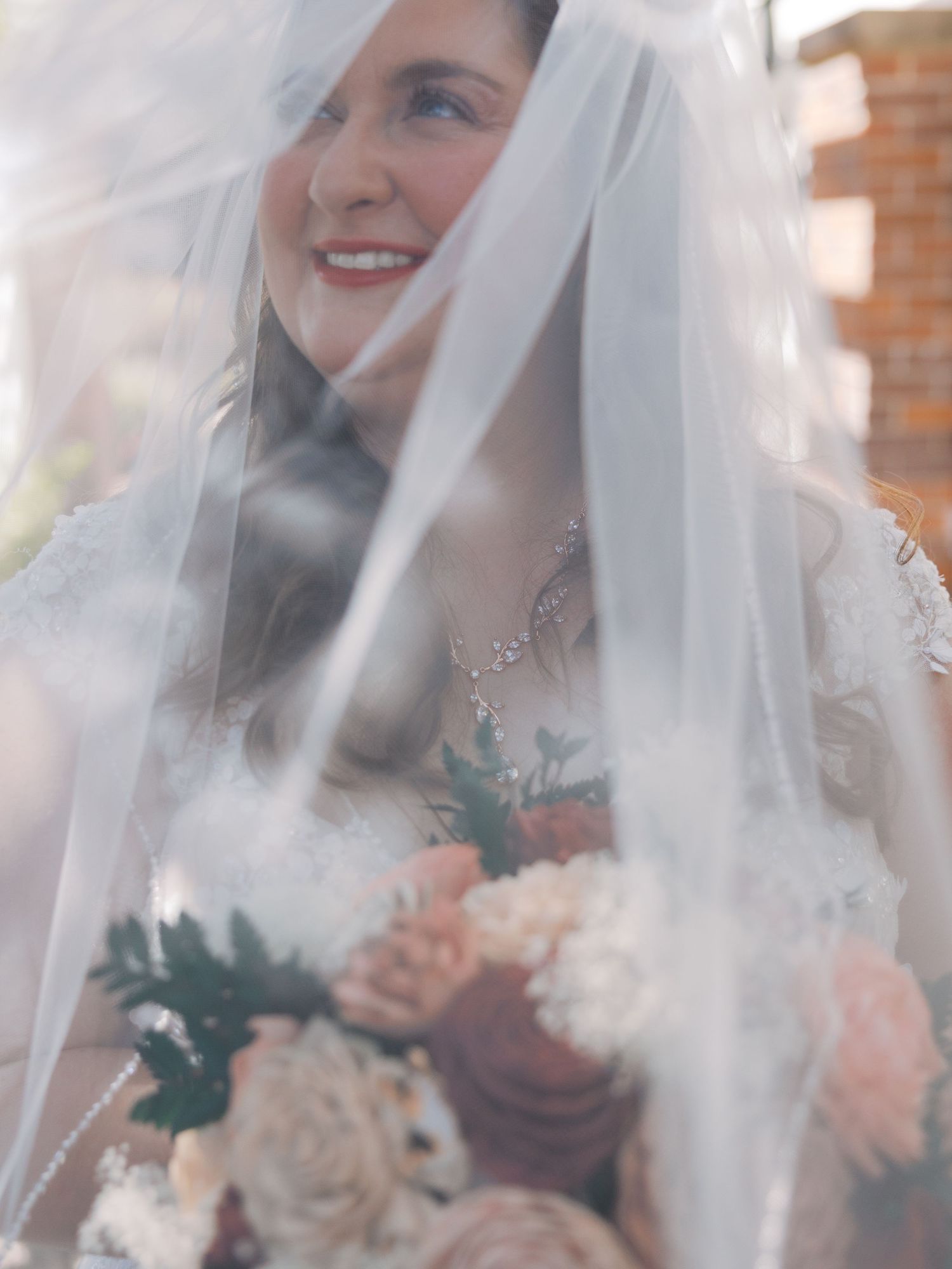 Smiling bride in lace gown holds blush bouquet under flowing veil outdoors on her wedding day.