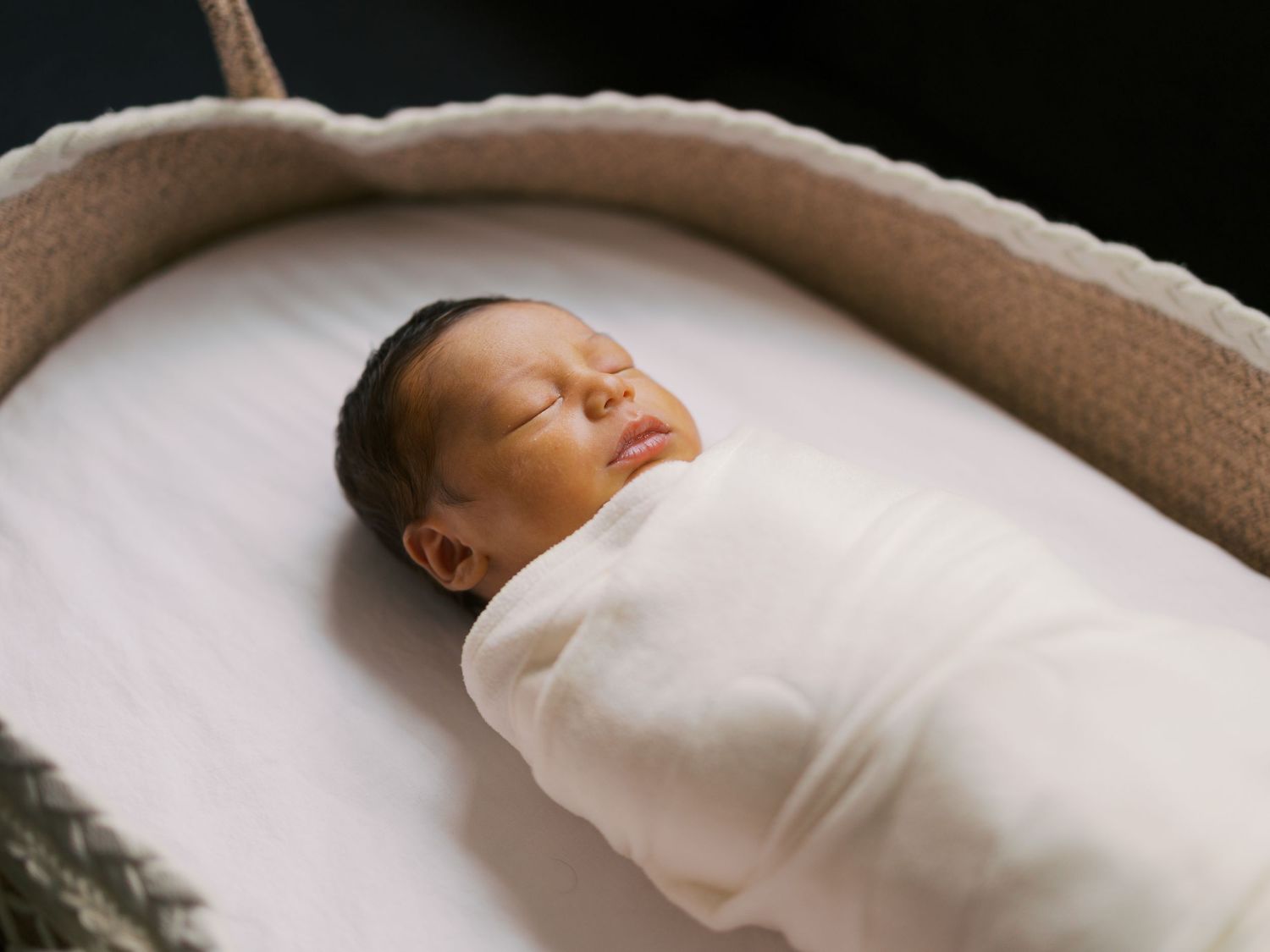 A peaceful newborn baby swaddled in a white blanket sleeping in a woven bassinet.