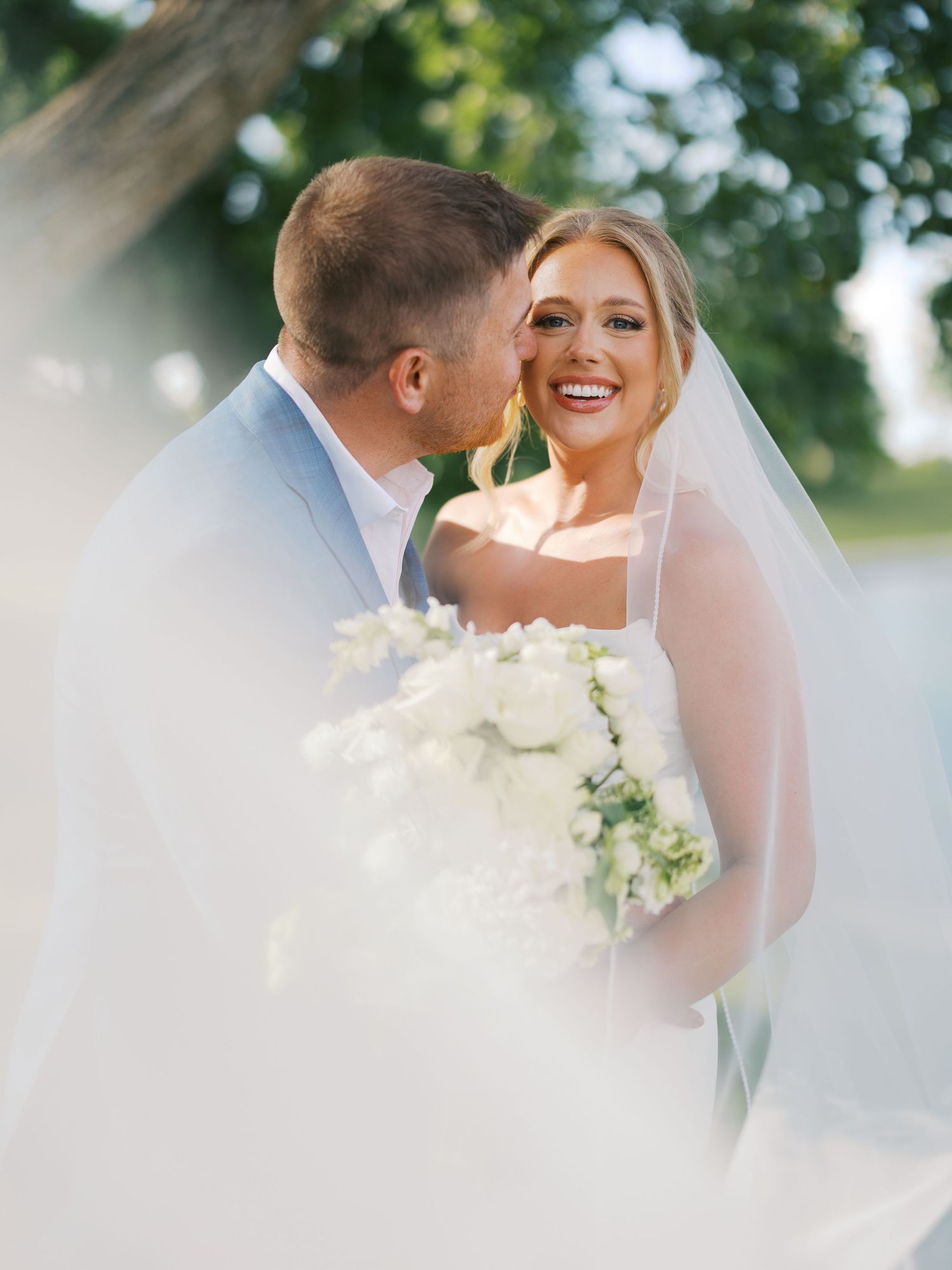Bride smiling joyfully as groom kisses her cheek outdoors, holding white floral bouquet, wearing wedding dress and veil.