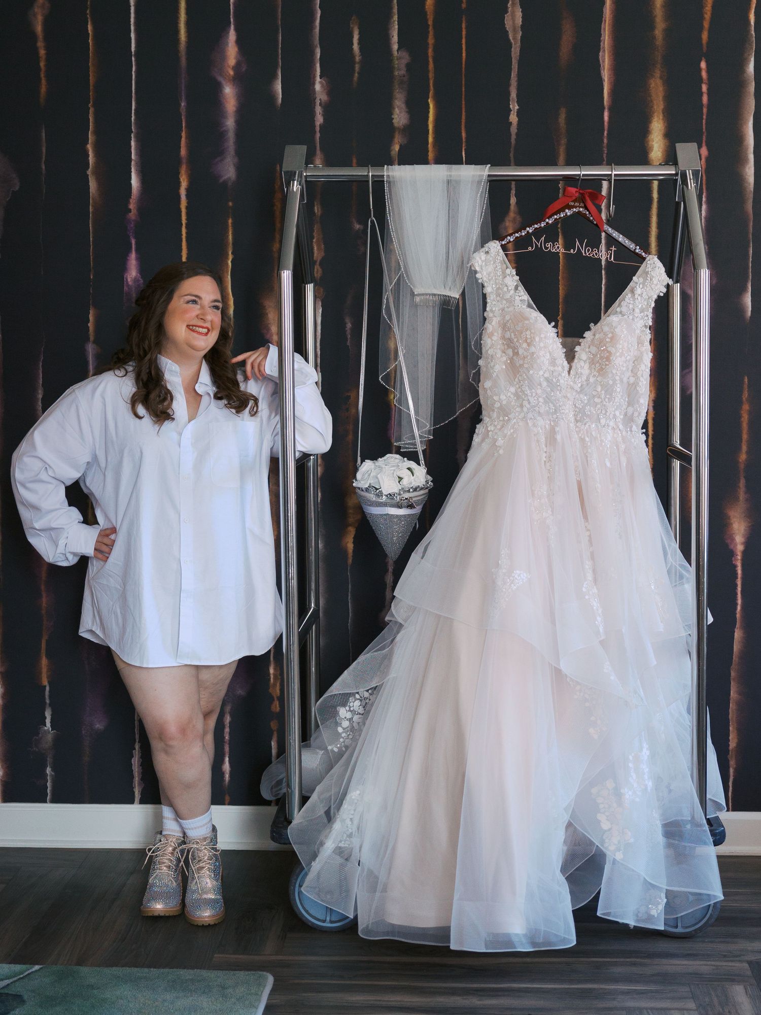 Woman in white shirt posing beside a elegant lace wedding dress displayed on a rack in a bridal shop.