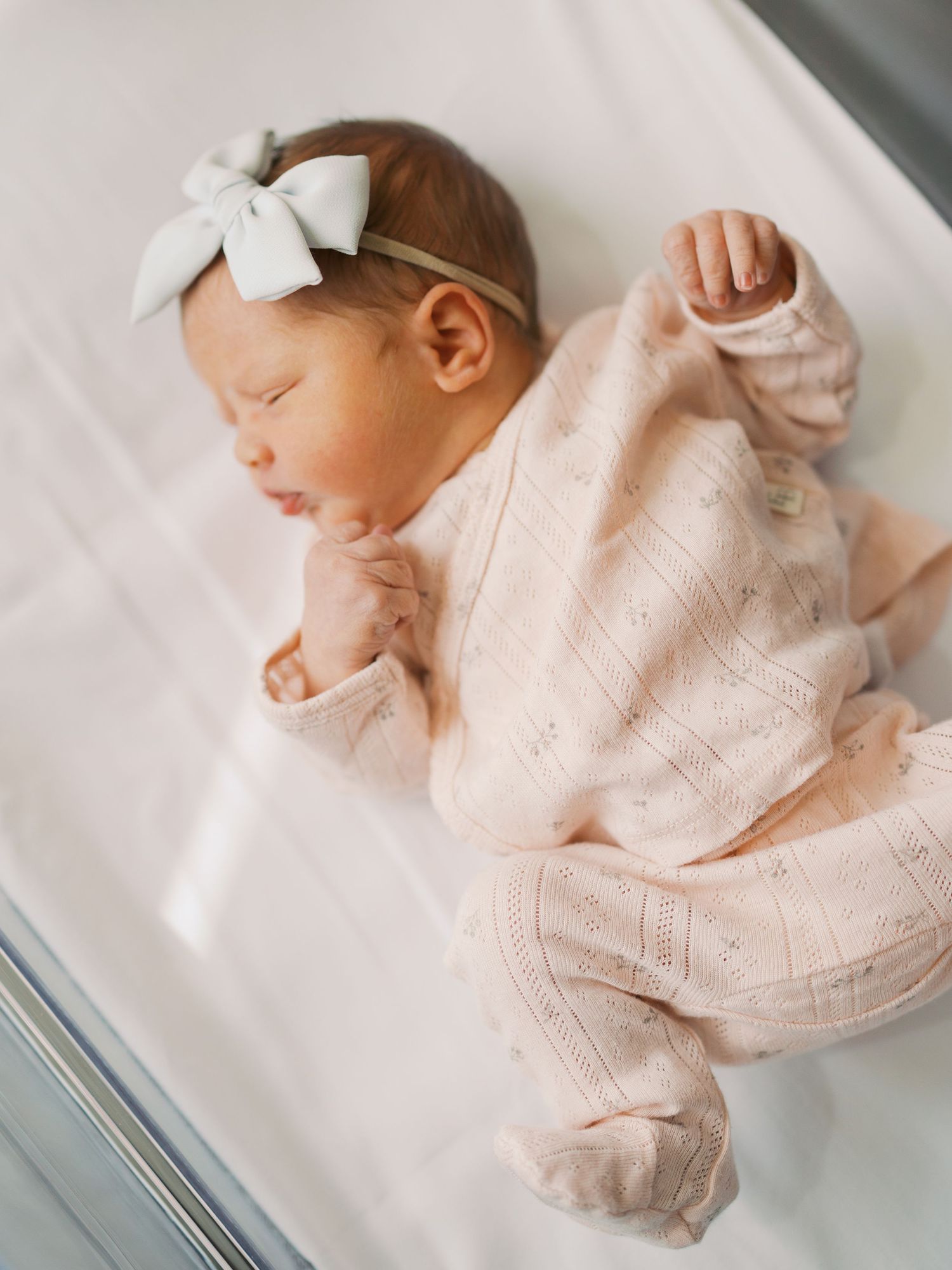 Sleeping newborn baby girl in pink knit outfit and white bow headband lying peacefully in hospital bassinet.