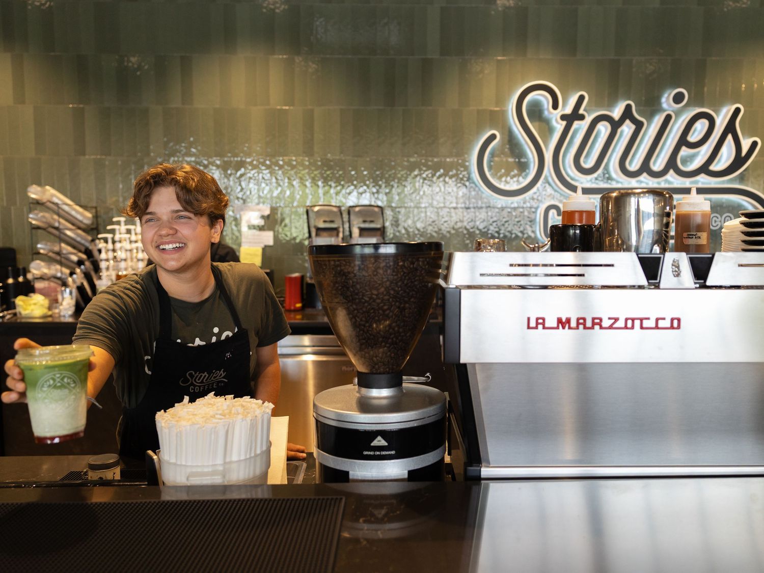 Smiling barista at La Marzocco espresso machine holding matcha drink at Stories coffee shop.
