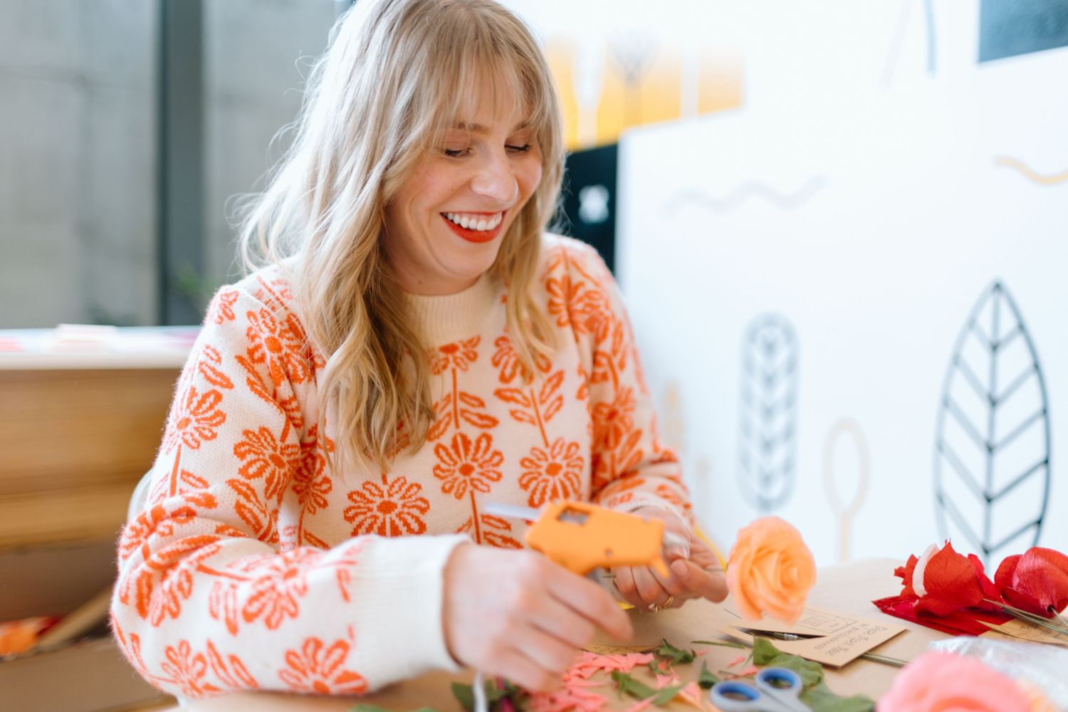 Smiling blonde woman in floral orange sweater crafting with colorful materials at a bright workspace.