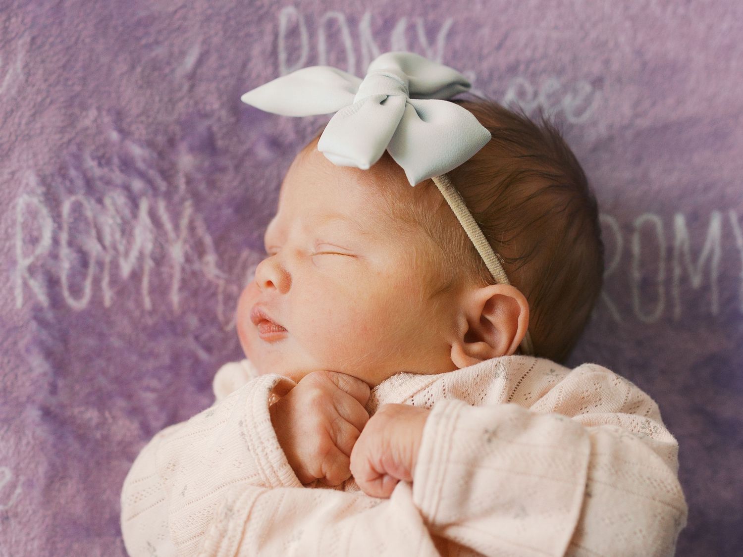 Newborn baby girl sleeping peacefully wearing a white bow headband on a purple blanket with the name Romy.