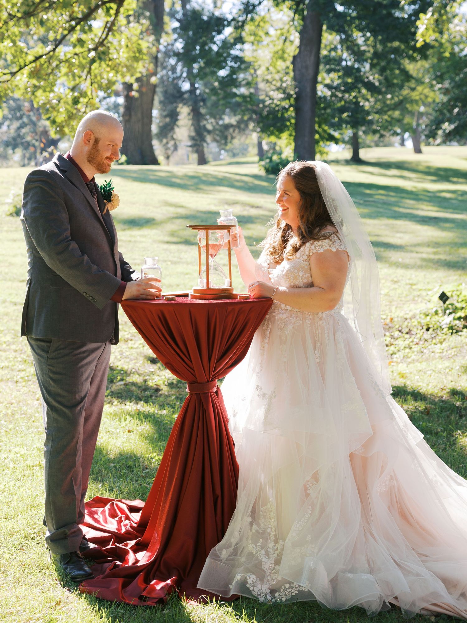 Bride and groom share a unity ceremony at a red draped table outdoors during a sunny wedding celebration.