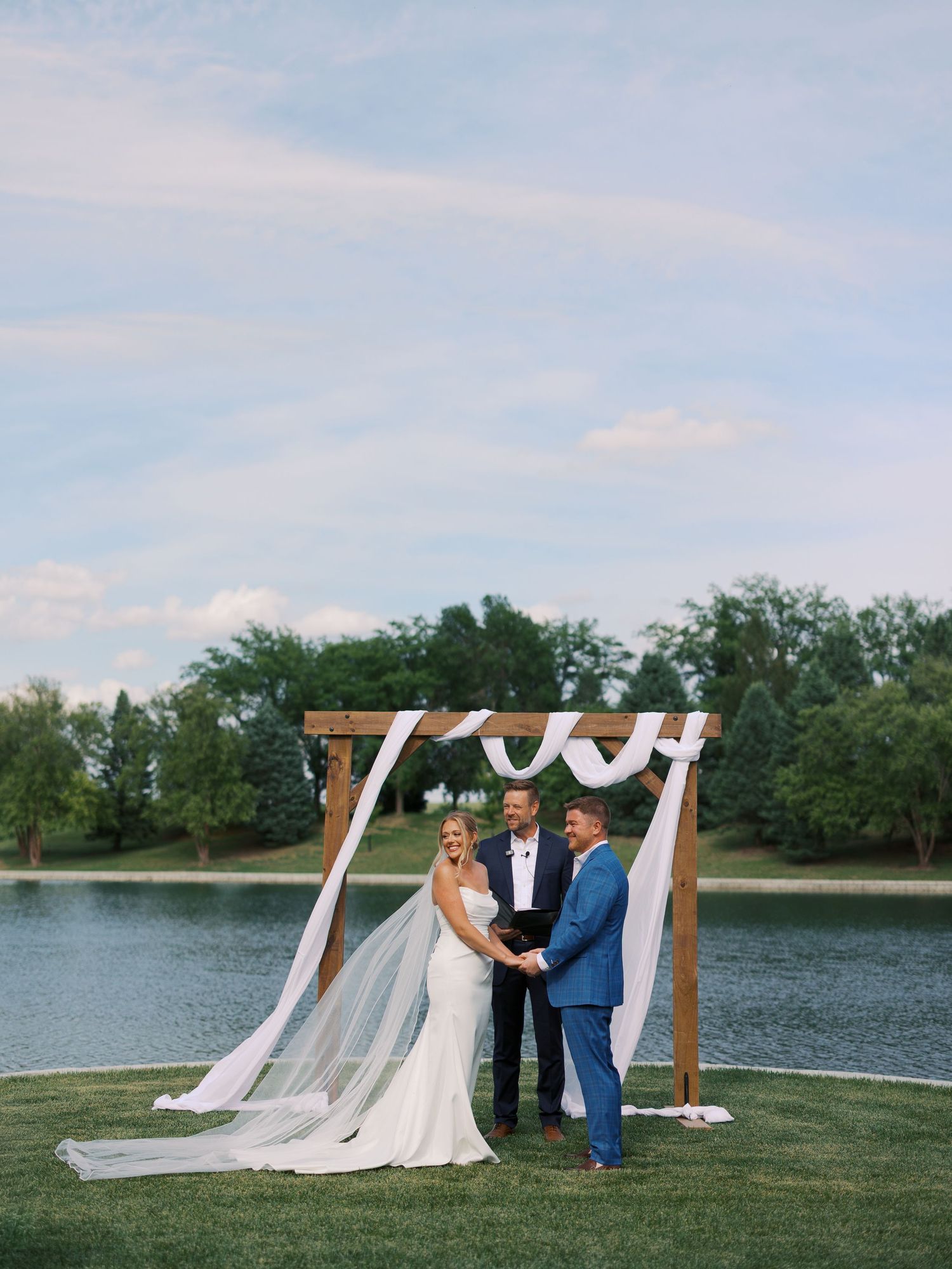 Bride and groom exchange vows under a wooden arch draped with white fabric beside a serene lake.